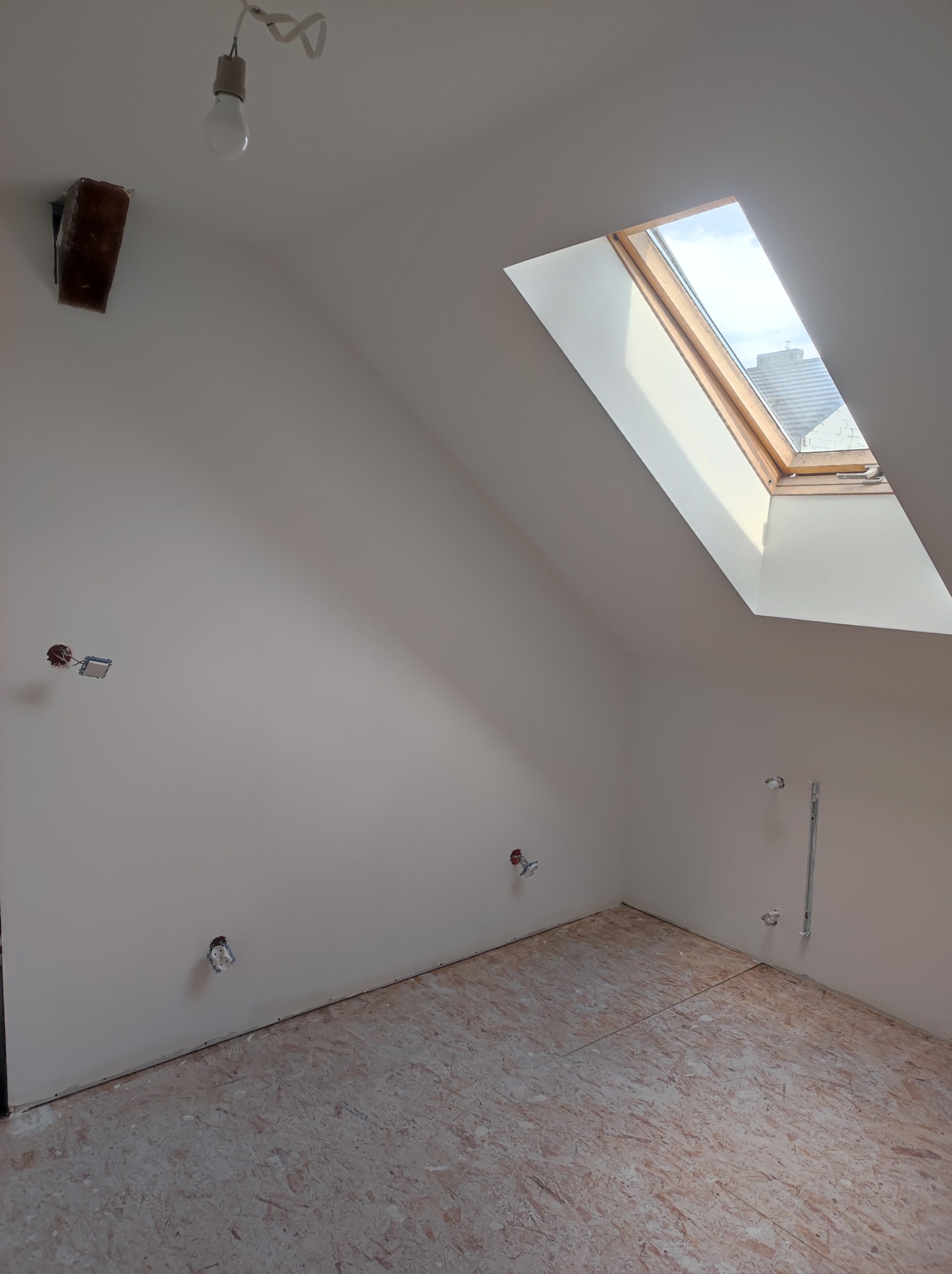 Poddasze po szpachlowaniu ścian i przygotowaniu podłogi z płyt OSB, z oknem dachowym i wystającymi kablami elektrycznymi i rurami hydraulicznymi, surowa żarówka zwisa z sufitu.