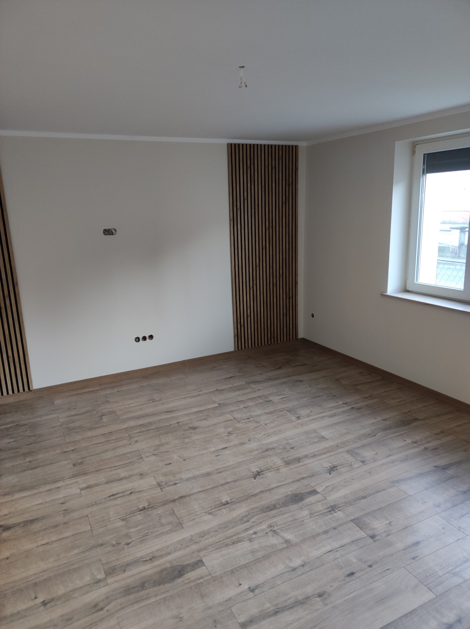 Pusty pokój po odświeżeniu: drewniane panele podłogowe, pionowe listwy dekoracyjne na ścianach, gładka biała ściana z widocznymi gniazdkami elektrycznymi, okno z roletą w białej ramie.