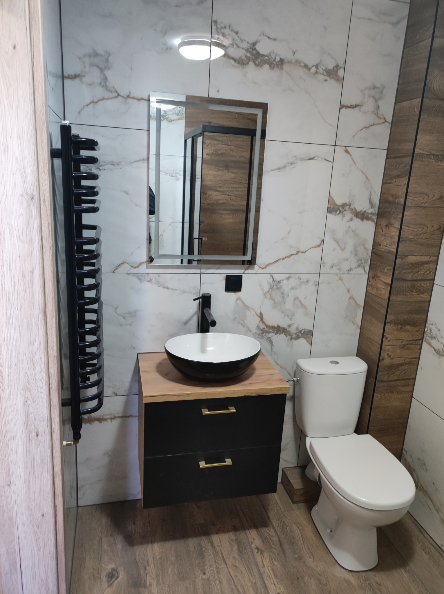 Nowoczesna łazienka z umywalką nablatową w czarnej misie, czarną armaturą, grzejnikiem drabinkowym w kolorze czarnym, lustrem z podświetleniem LED, płytkami imitującymi marmur oraz drewnopodobnymi...