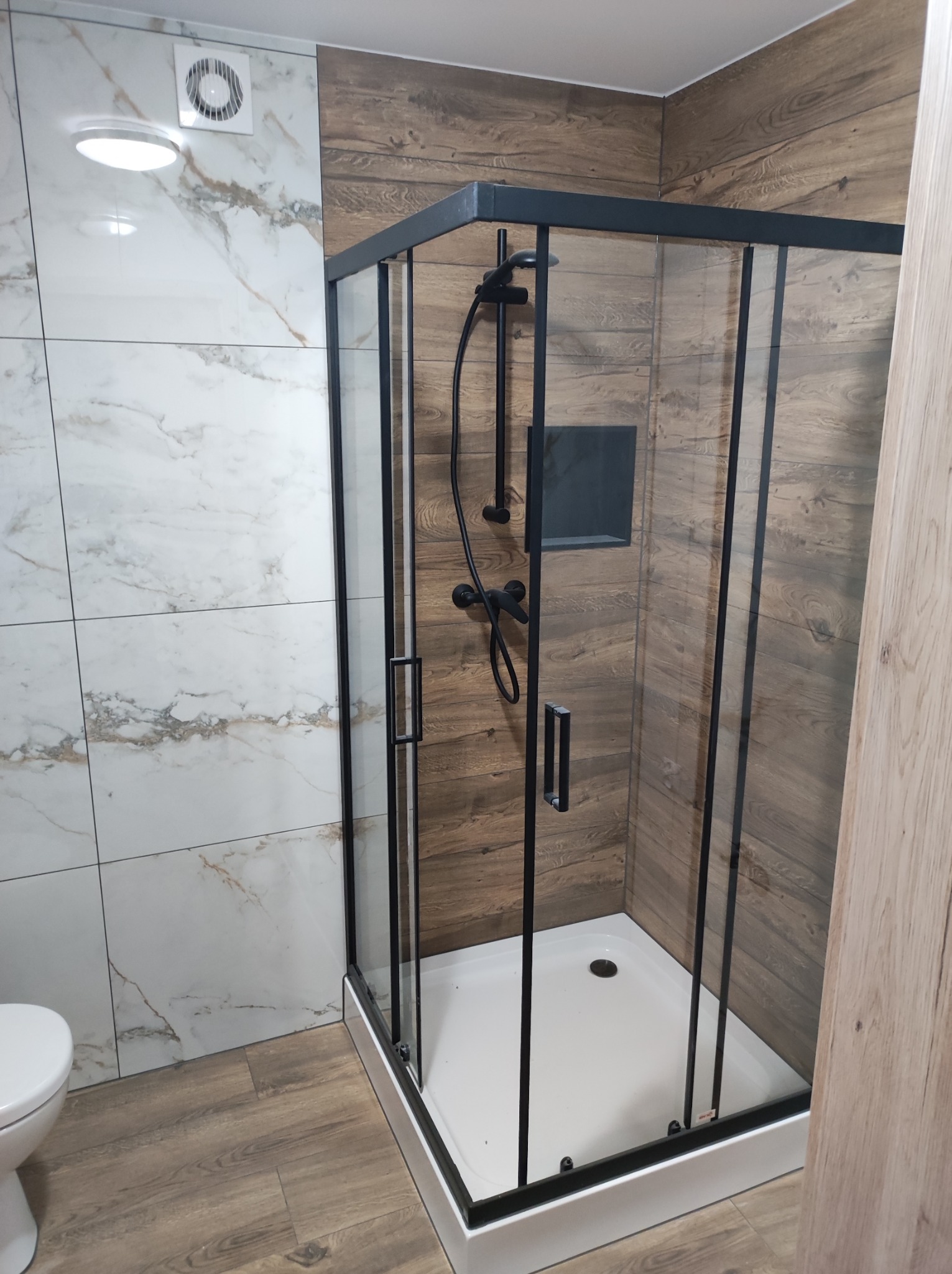 Nowoczesna kabina prysznicowa z czarnymi profilami i armaturą, umieszczona w łazience z płytkami imitującymi marmur i drewno, widoczny fragment toalety po lewej stronie.