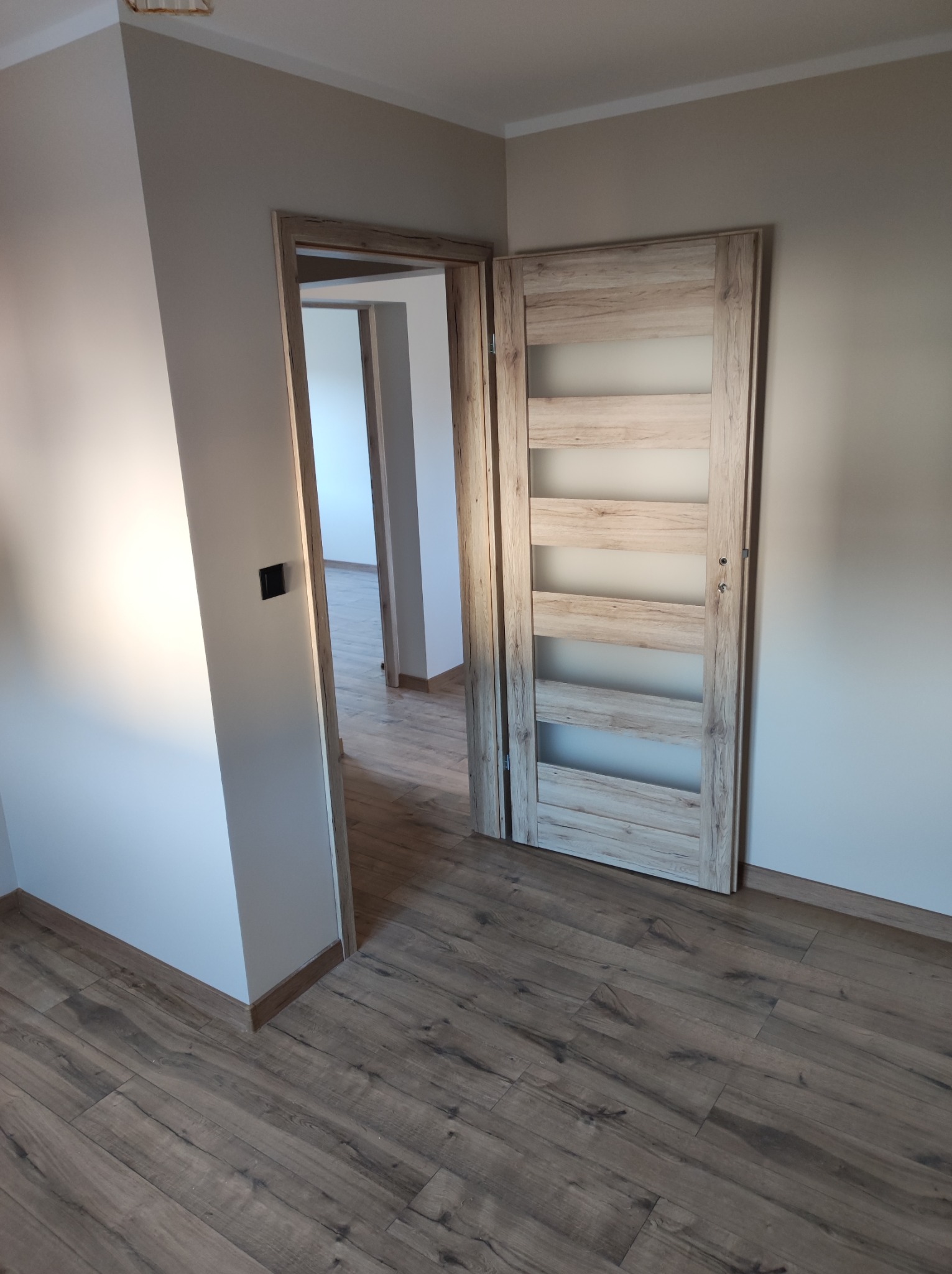 Zamknięte drzwi wewnętrzne z drewnianą ramą i przeszkleniami, widoczne na tle świeżo wykończonego wnętrza z panelami podłogowymi w odcieniach brązu i beżowe ściany w nowym budynku.