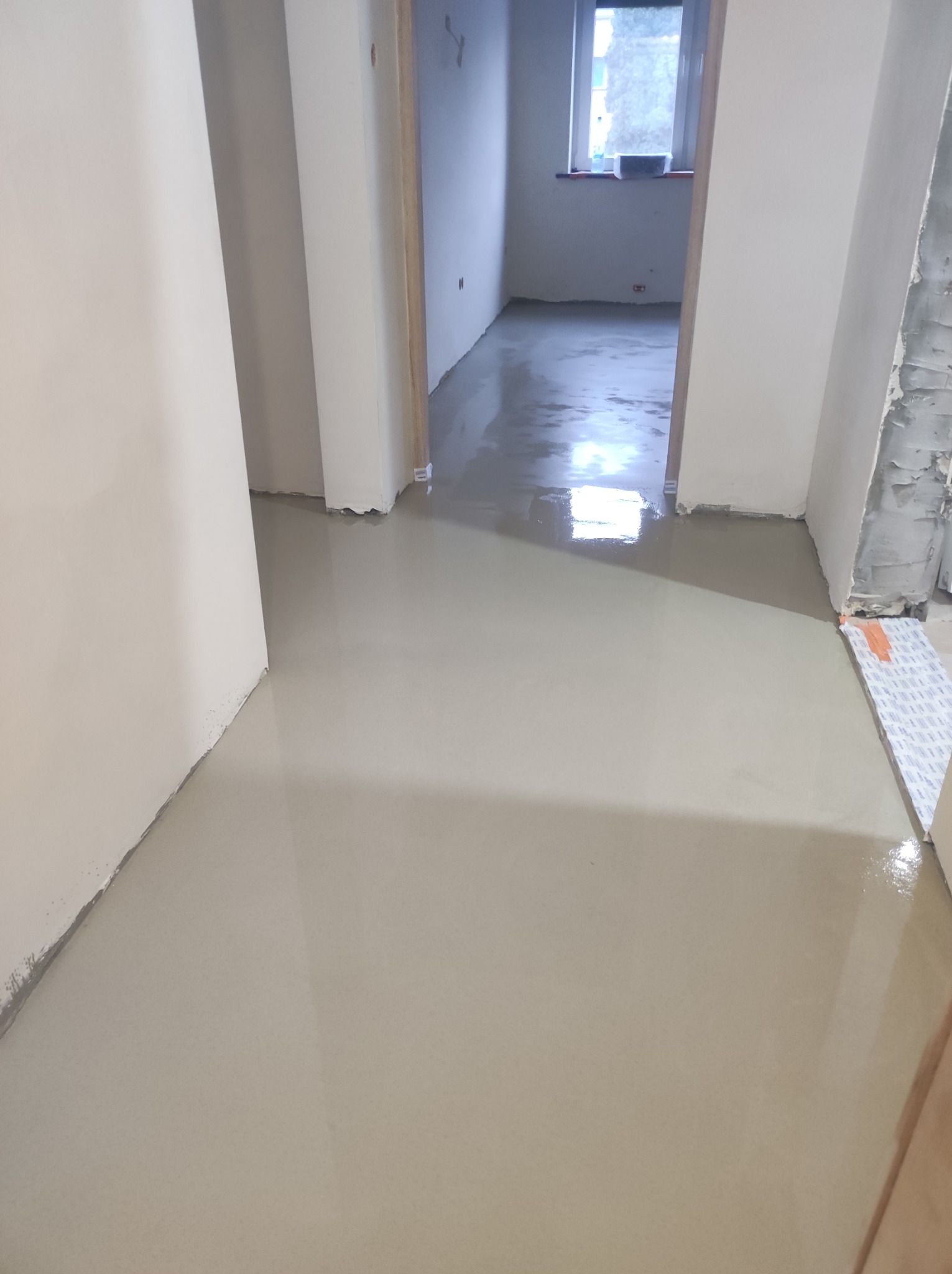 Wylany, mokry podkład podłogowy w jasnym odcieniu szarości, widoczne ściany i fragmenty ościeżnic drzwiowych w trakcie prac wykończeniowych.