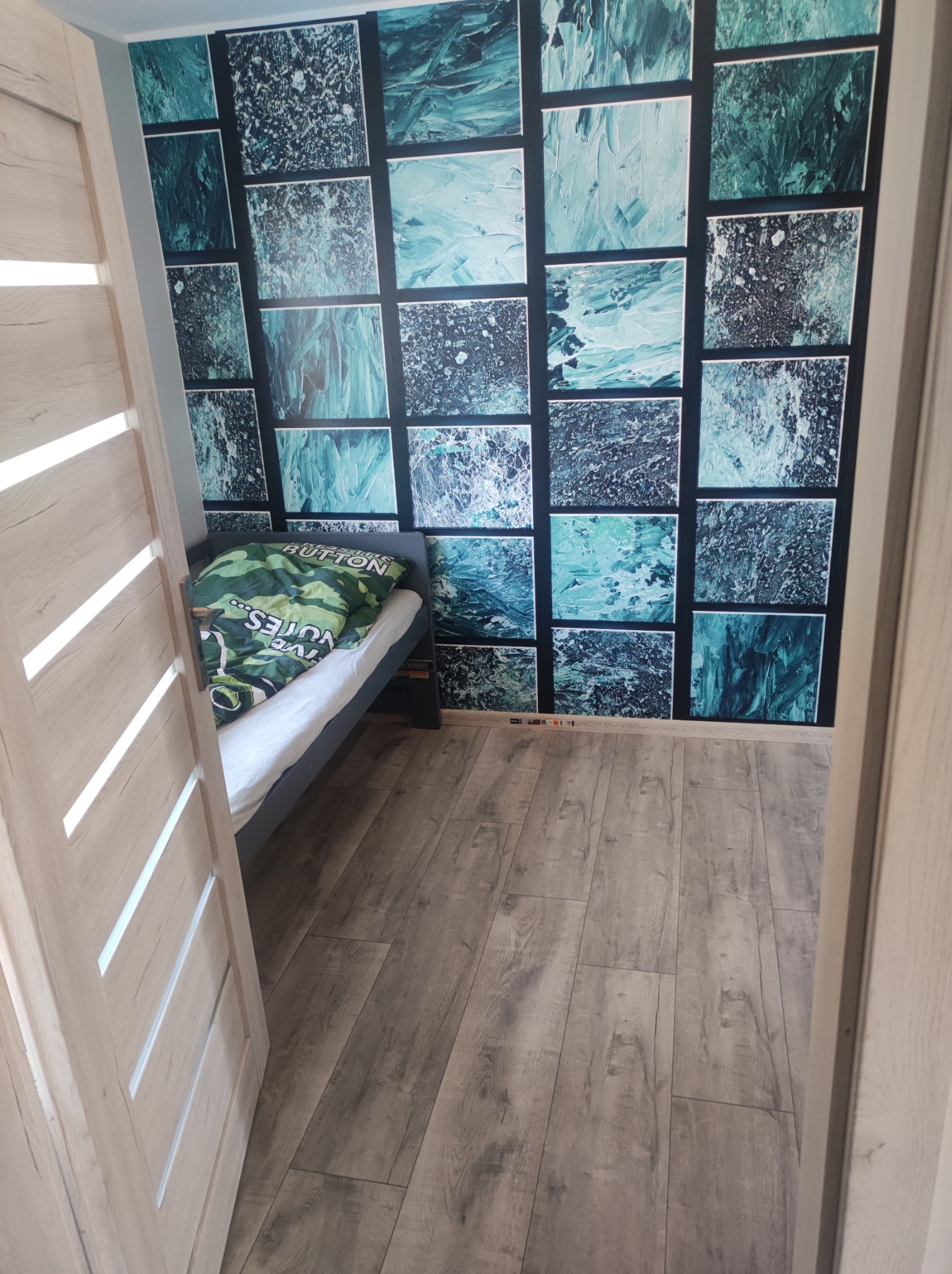 Pokój z łóżkiem, drewnianą podłogą i ścianą pokrytą tapetą w geometryczne wzory, widok zza uchylonych, jasnych drzwi.