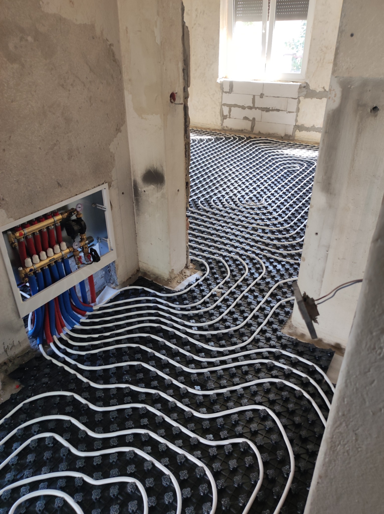 Rozdzielacz ogrzewania podłogowego z czerwonymi i niebieskimi rurami oraz białe rurki grzewcze ułożone w pętle na czarnej macie izolacyjnej w surowym wnętrzu.