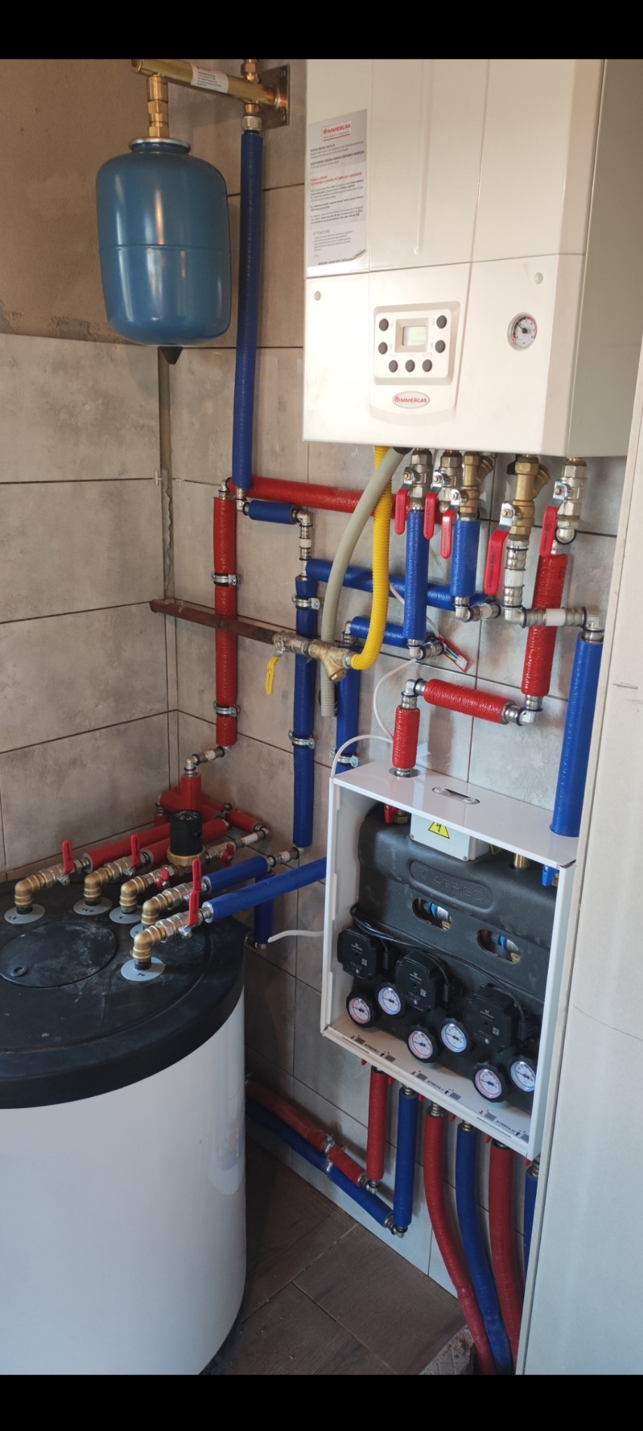 Złożona instalacja grzewcza z kotłem Immergas, zbiornikiem akumulacyjnym i rozdzielaczem w pomieszczeniu z szarymi płytkami.