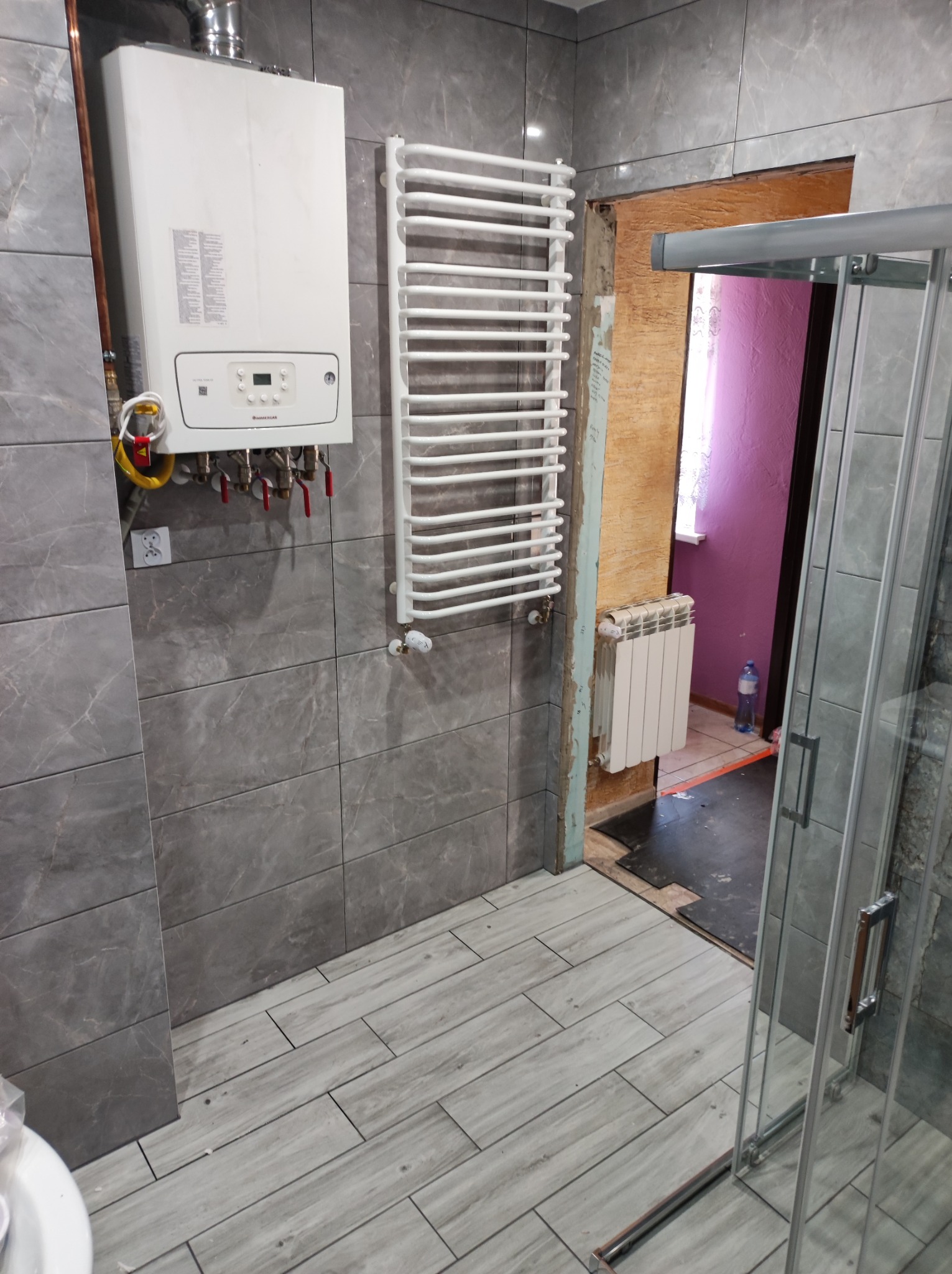 Szare płytki imitujące marmur na ścianie łazienki, biały grzejnik drabinkowy, piec gazowy i fragment kabiny prysznicowej w trakcie remontu, widoczne niedokończone obramowanie drzwi do innego...