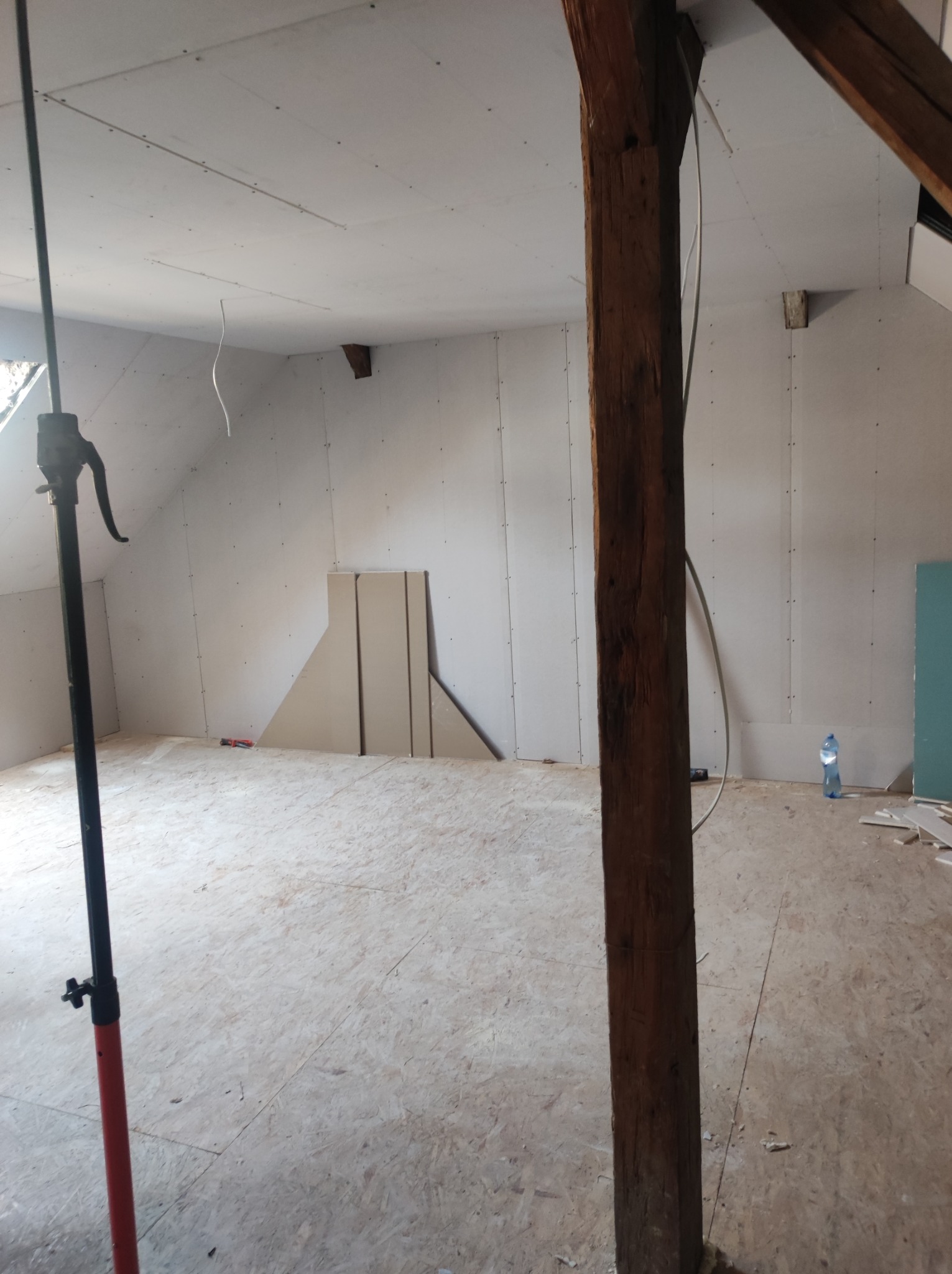 Poddasze w trakcie adaptacji, ściany wyłożone płytami gipsowo-kartonowymi, widoczny drewniany słup konstrukcyjny, podłoga z płyt OSB, narzędzia i materiały budowlane.