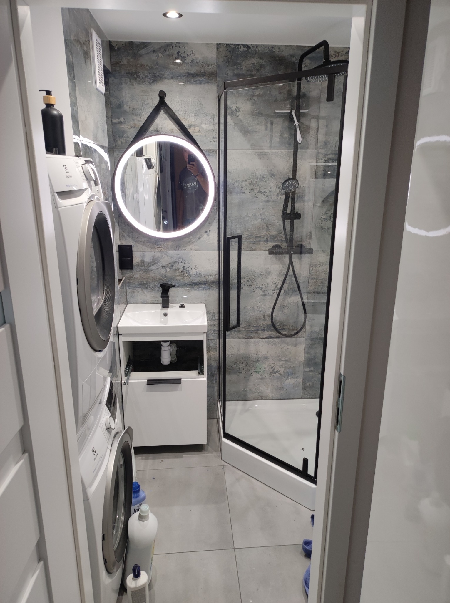 Nowoczesna łazienka z kabiną prysznicową typu walk-in, umywalką z szafką, okrągłym lustrem z podświetleniem LED i pralką Electrolux ustawioną obok, widoczne szare płytki imitujące kamień...