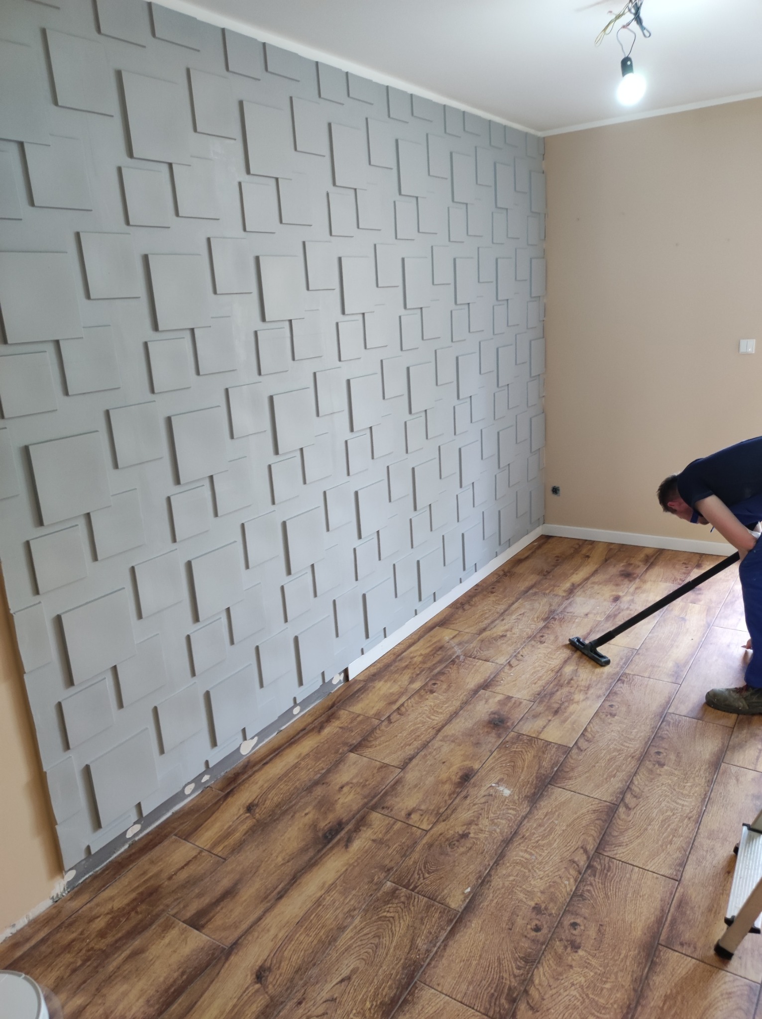 Montaż paneli podłogowych w pomieszczeniu z dekoracyjną ścianą z paneli 3D; widoczne narzędzie do czyszczenia podłogi i drabina