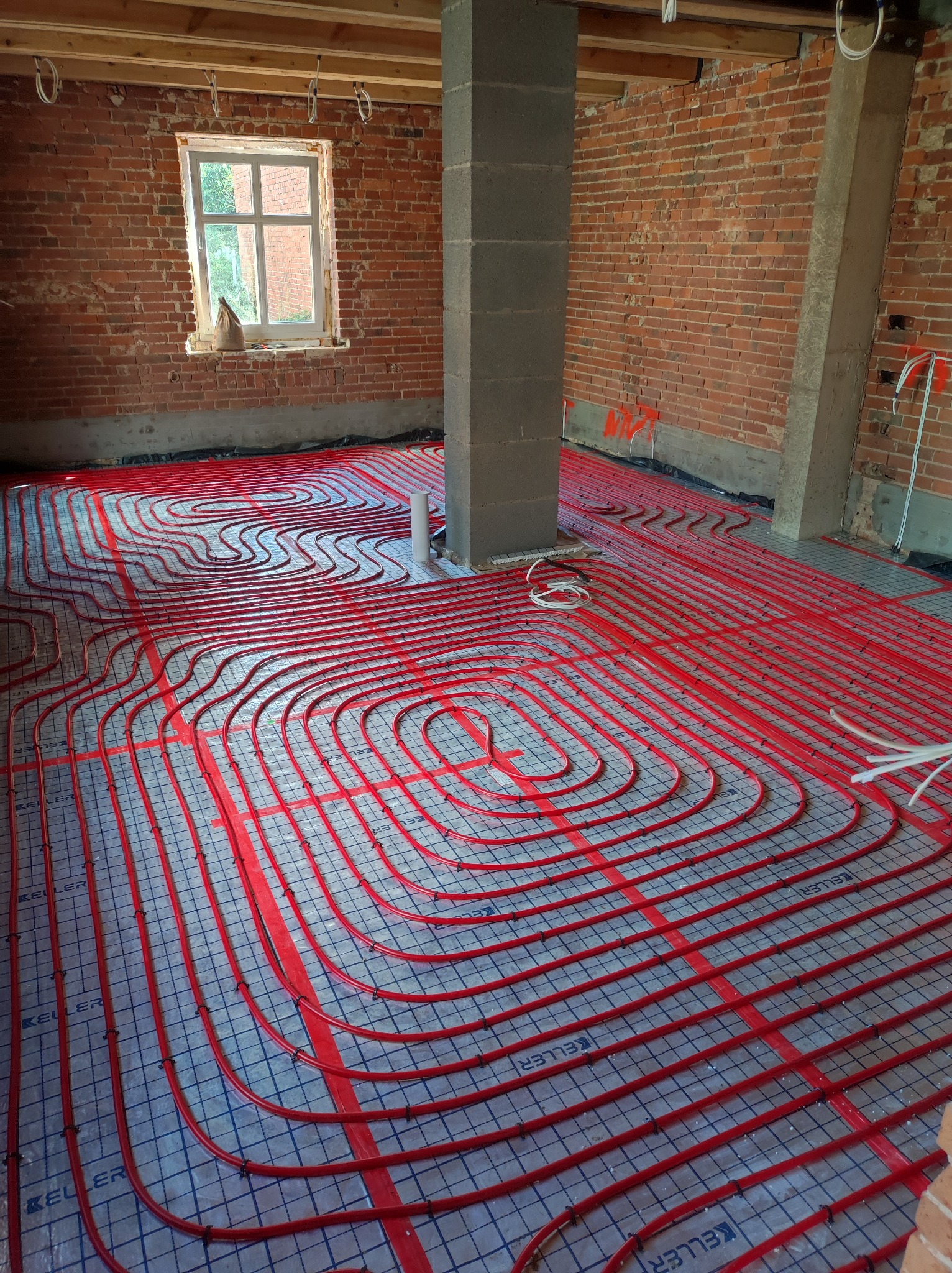 Czerwone rury ogrzewania podłogowego ułożone na folii w surowym wnętrzu z ceglanymi ścianami i betonowym słupem. Widoczne okno z workiem cementu na parapecie.