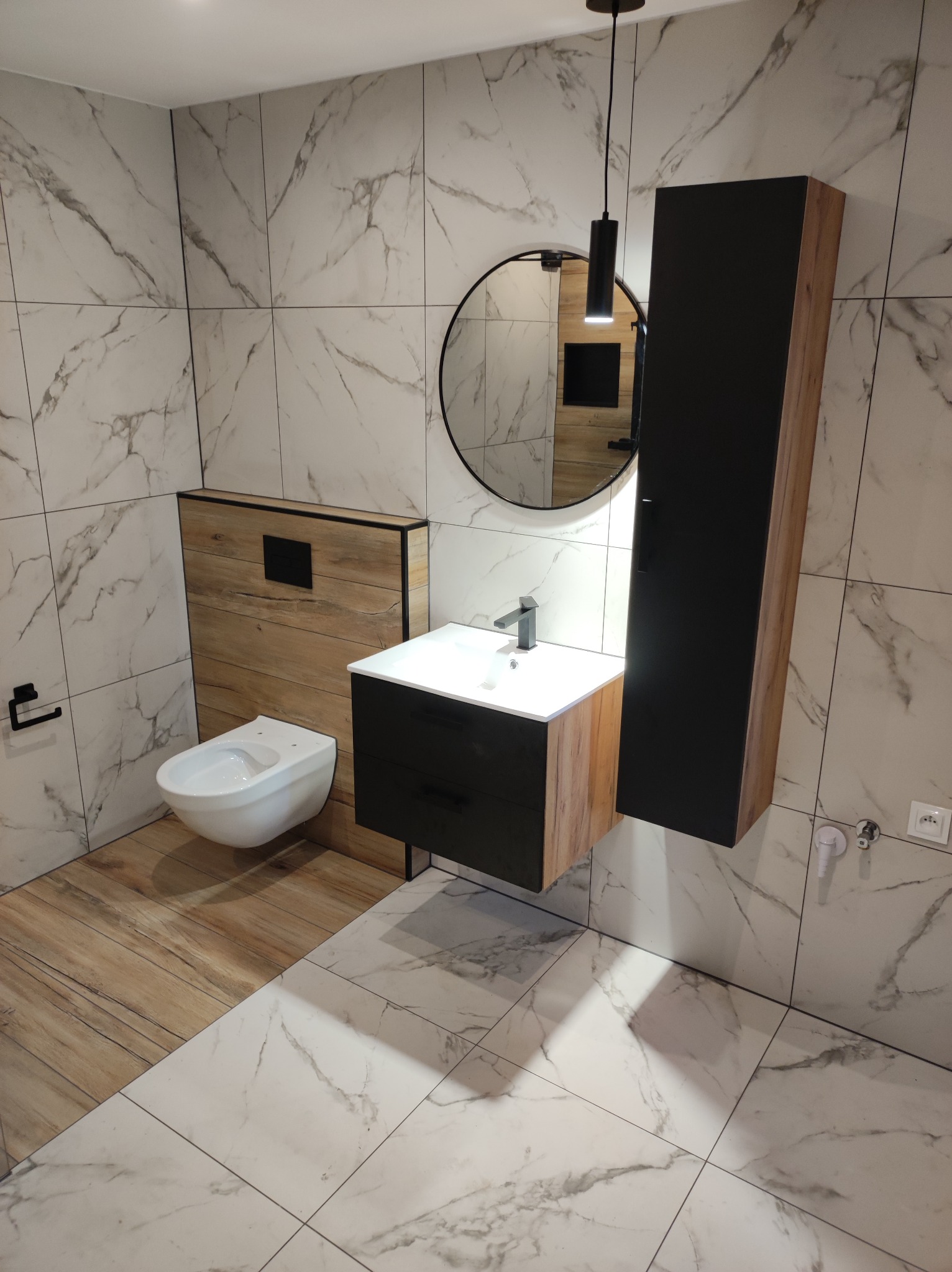 Nowoczesna łazienka z marmurowymi płytkami, okrągłym lustrem i wiszącą szafką. Uwagę zwraca kontrast faktur drewna i czerni w zabudowie toalety i umywalki.