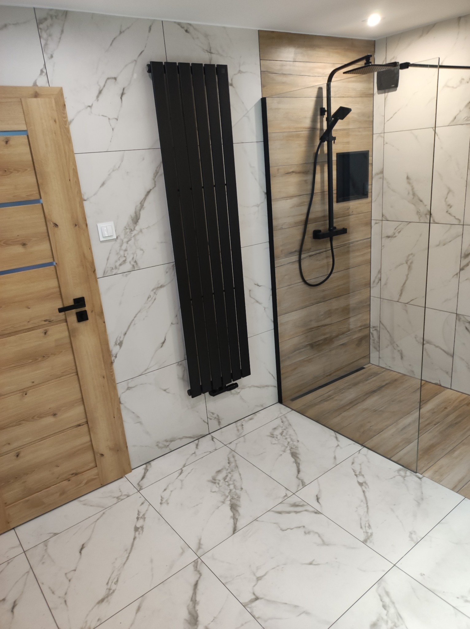 Nowoczesna łazienka z kabiną typu walk-in, grzejnikiem drabinkowym i marmurowymi płytkami. Drewniane akcenty na drzwiach i ścianie prysznica.