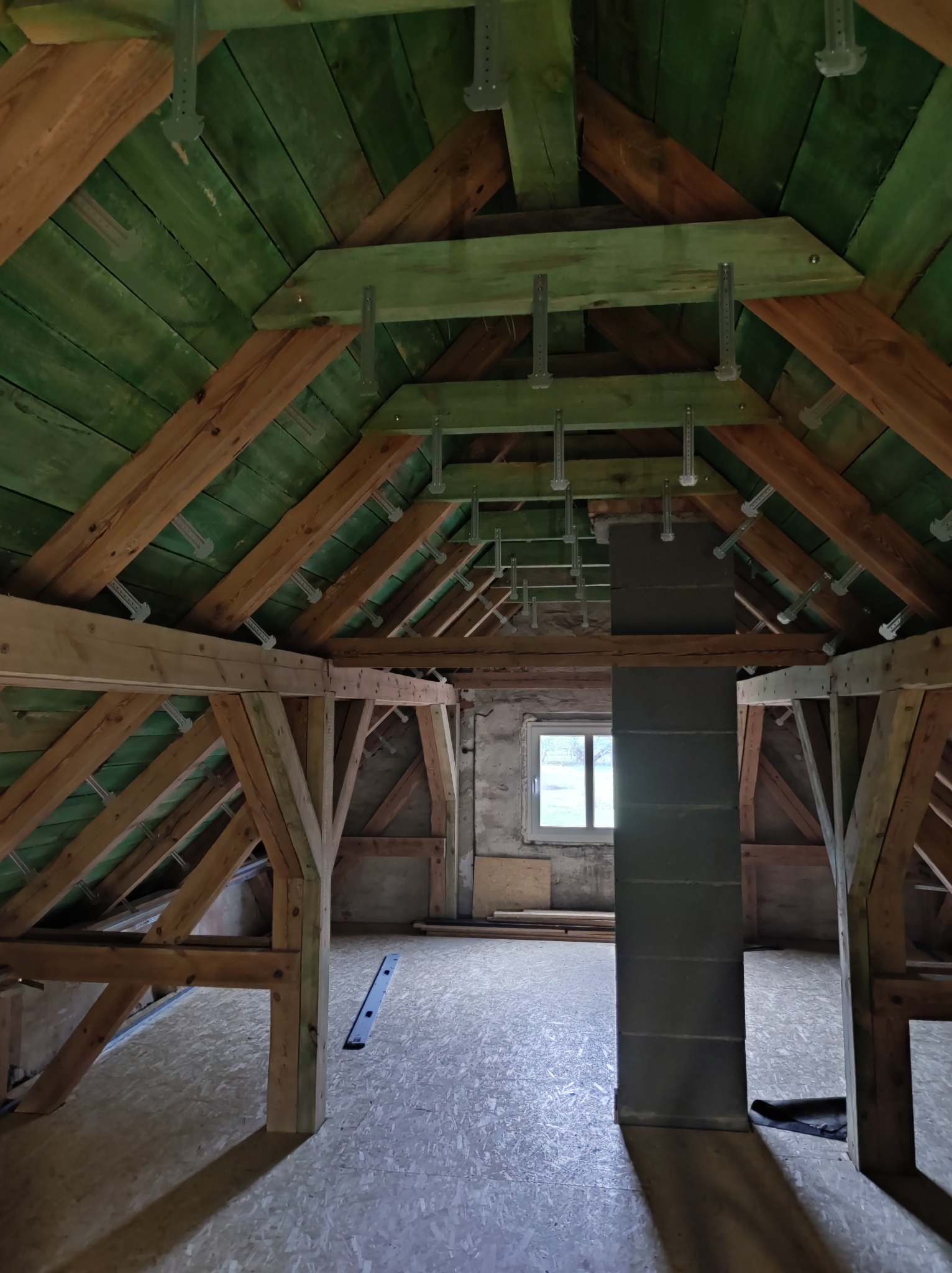 Adaptacja poddasza: drewniana konstrukcja dachu z zieloną boazerią, widoczne elementy mocujące, podłoga z płyt OSB, okno w tle. Etap prac wykończeniowych.