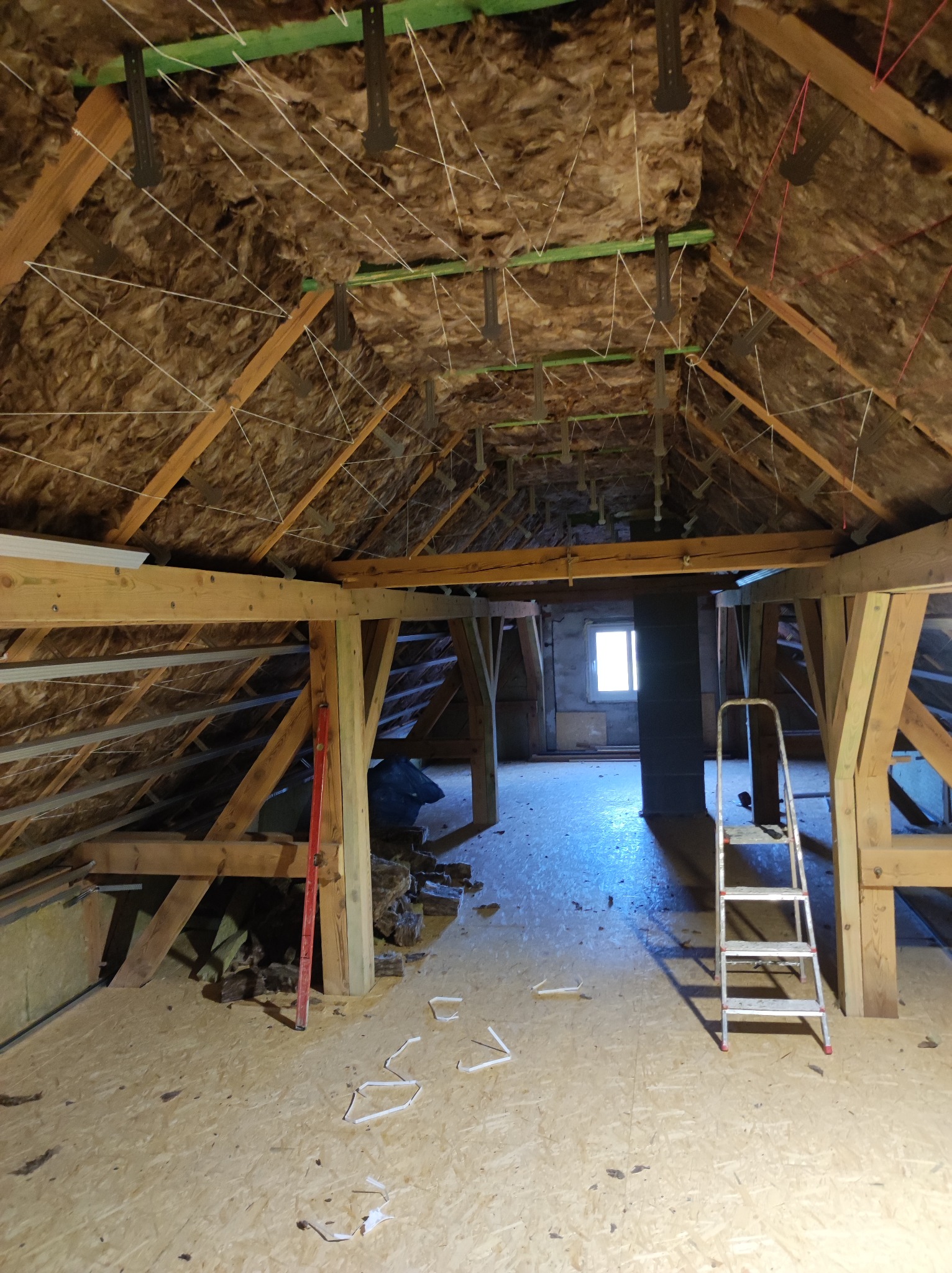 Ocieplenie poddasza wełną mineralną, widoczne drewniane krokwie, metalowe wieszaki i sznurki. Częściowo ułożona podłoga z płyt OSB. Prace w toku.