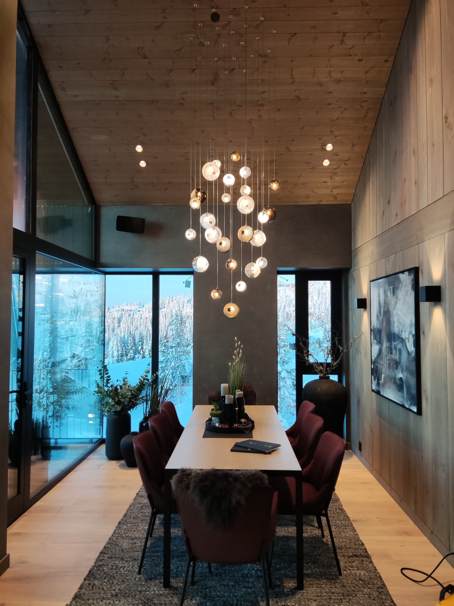 Jasne wnętrze jadalni w domu szkieletowym z drewnianym sufitem i ścianami, panoramiczne okno z widokiem na ośnieżony krajobraz, nowoczesny żyrandol z kulistymi kloszami nad stołem z krzesłami...