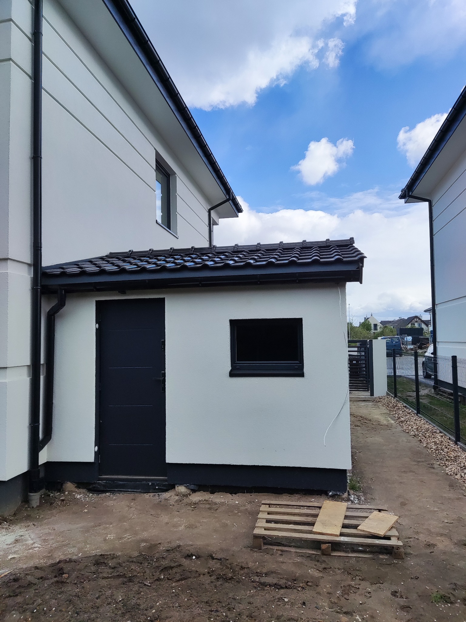 Nowoczesny, biały dom szkieletowy z ciemną stolarką okienną i drzwiami, widok na fragment elewacji i otoczenie w trakcie prac wykończeniowych, z paletą leżącą na ziemi.