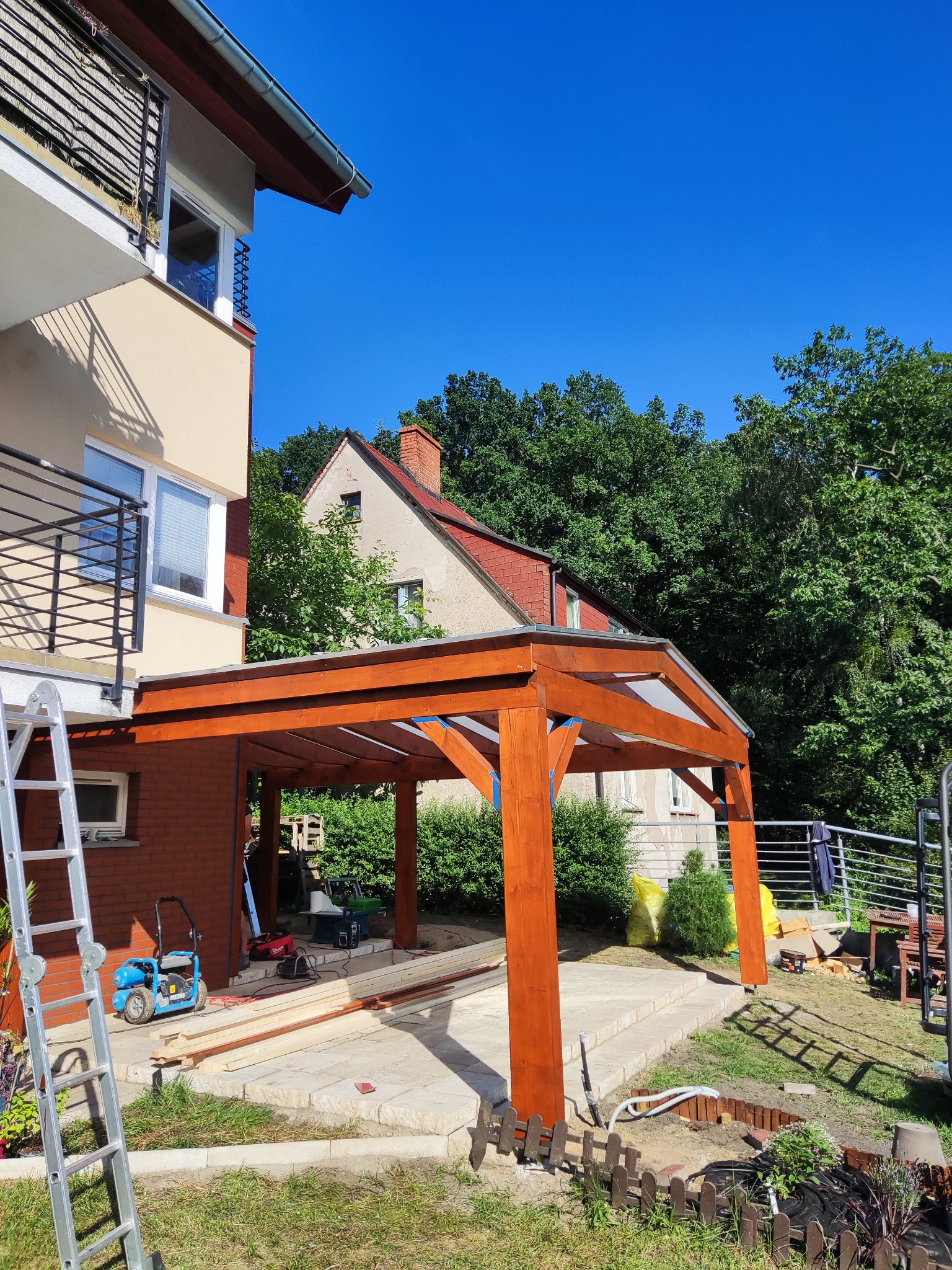 Konstrukcja z drewna w kolorze miodowym, prawdopodobnie zadaszenie tarasu lub carport, w trakcie budowy, widoczne narzędzia i materiały budowlane, dom w tle.