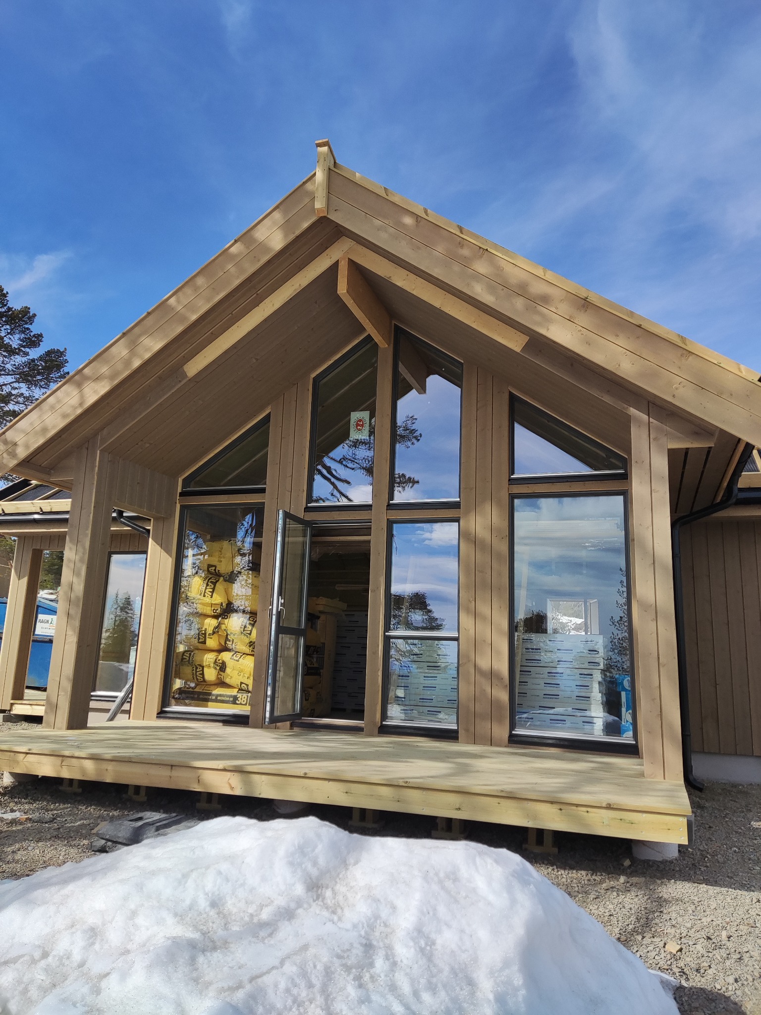 Surowy, drewniany dom w konstrukcji szkieletowej z dużymi oknami, częściowo otwartymi drzwiami i drewnianym tarasem, otoczony topniejącym śniegiem.