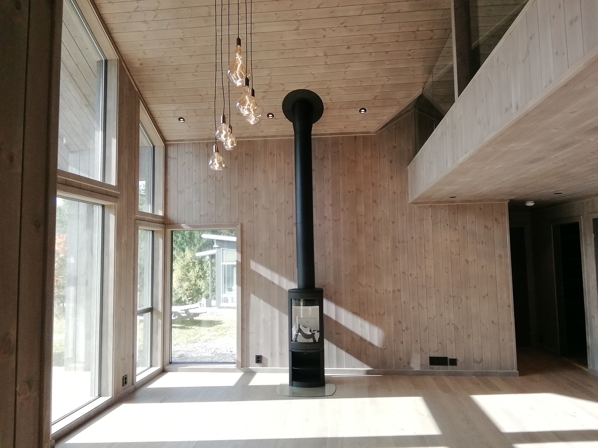 Wnętrze domu z drewnianą boazerią, dużymi oknami i czarnym piecem wolnostojącym z rurą kominową, wiszące żarówki dekoracyjne