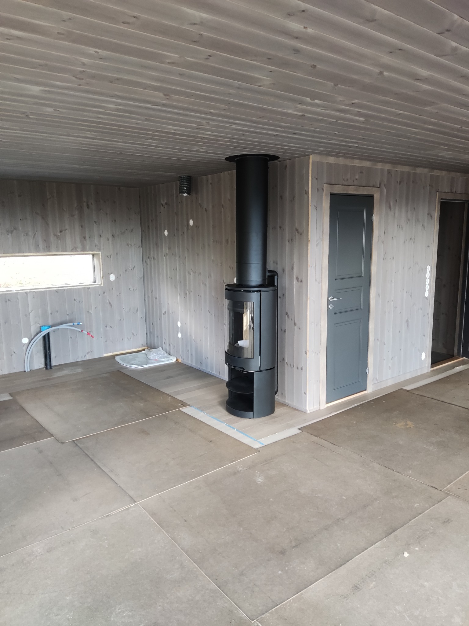 Wnętrze domu w trakcie budowy z drewnianą boazerią, czarnym piecem wolnostojącym, przygotowaną podłogą i widocznymi instalacjami.