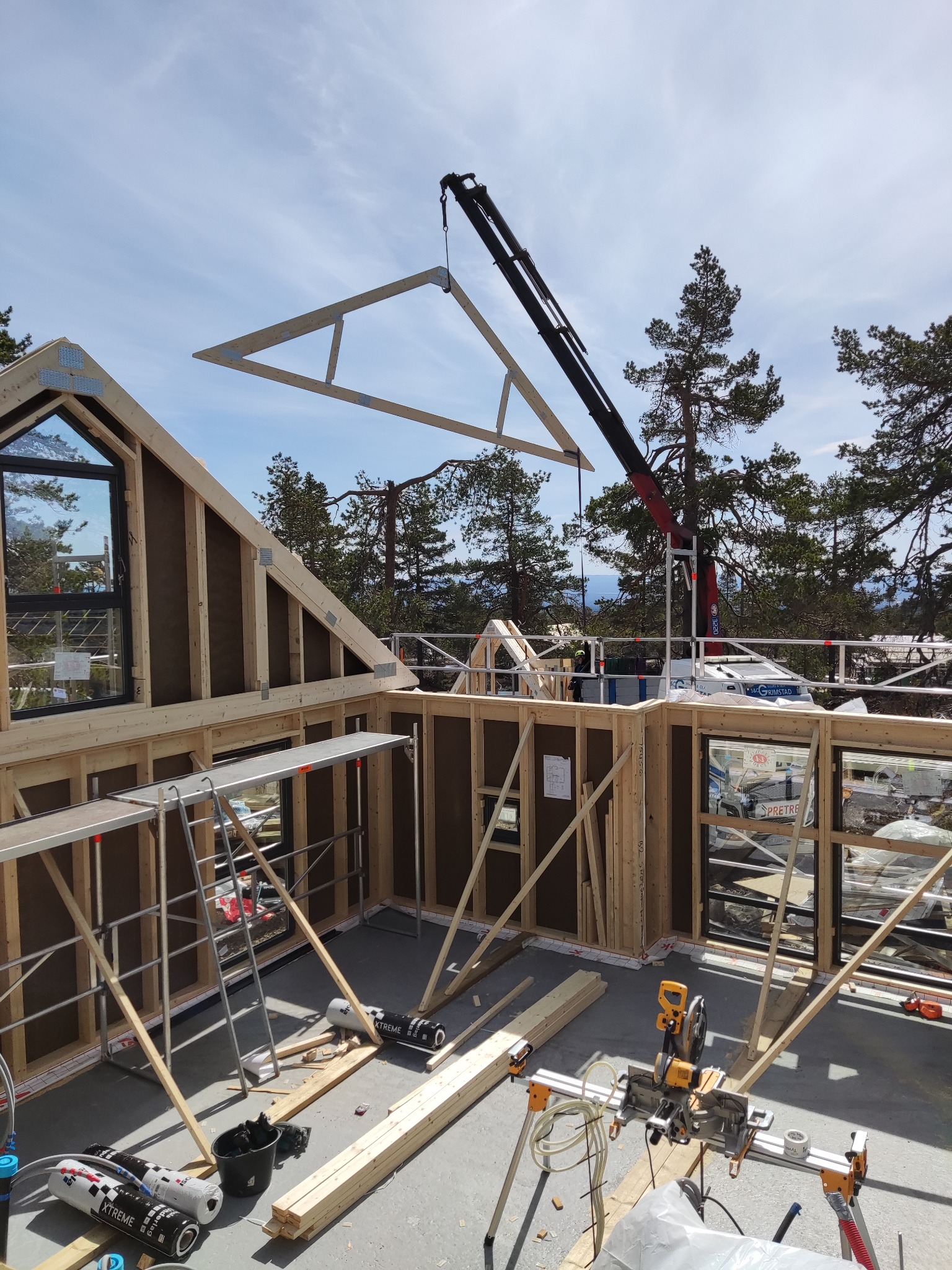 Montaż drewnianej konstrukcji domu szkieletowego z użyciem dźwigu, widoczne rusztowania, narzędzia i elementy konstrukcyjne.