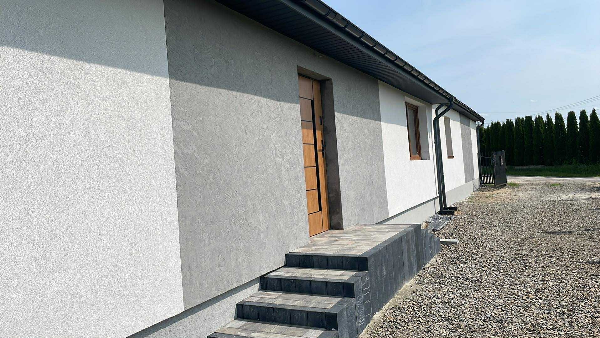 Elewacja domu jednorodzinnego z dekoracyjnym tynkiem w odcieniach szarości i bieli. Widoczne schody wejściowe i fragment ogrodu.