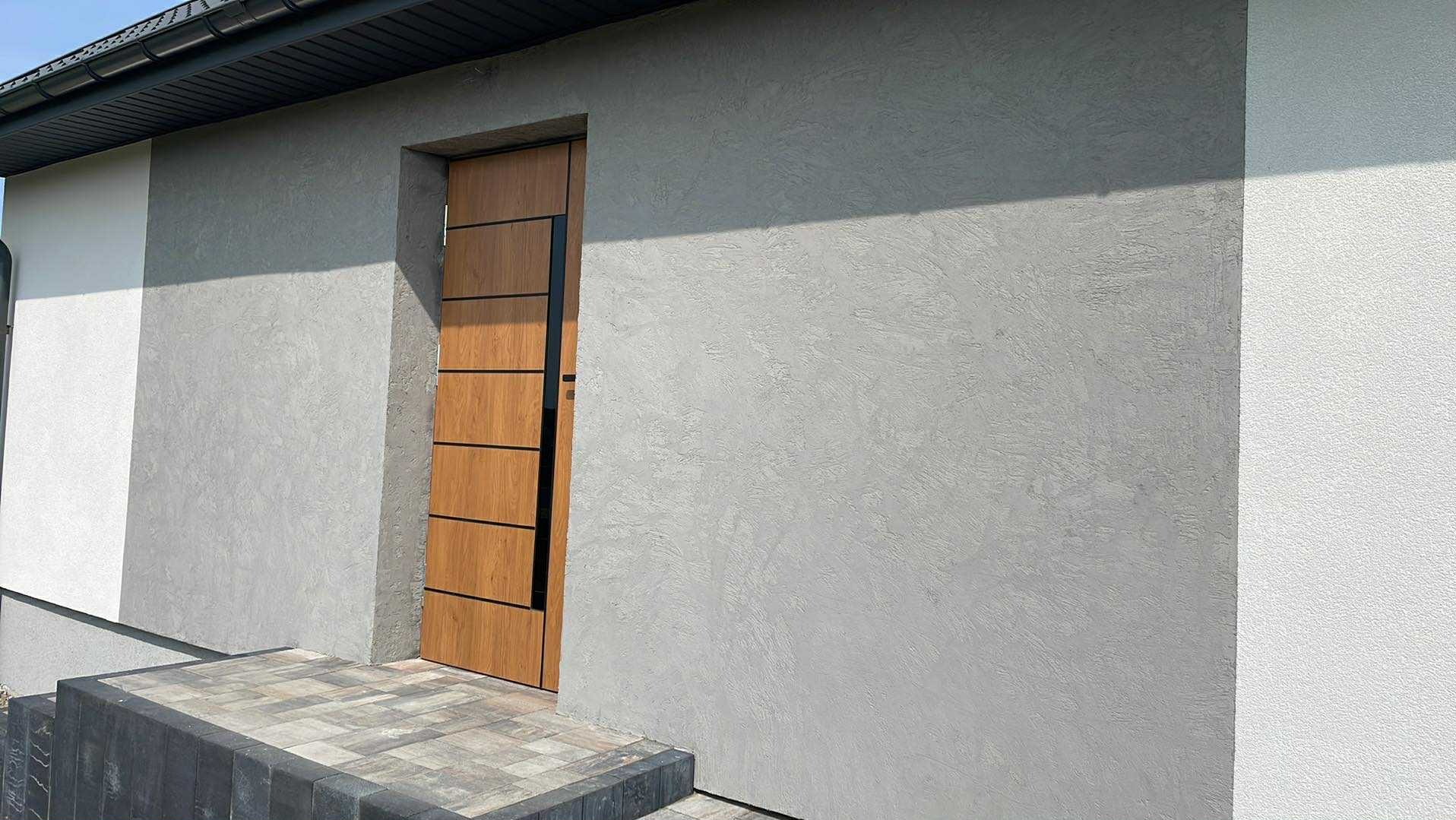 Elewacja domu z szarą, dekoracyjną strukturą tynku i nowoczesnymi drzwiami wejściowymi z okładziną imitującą drewno. Widoczne schody wejściowe z kostki brukowej.