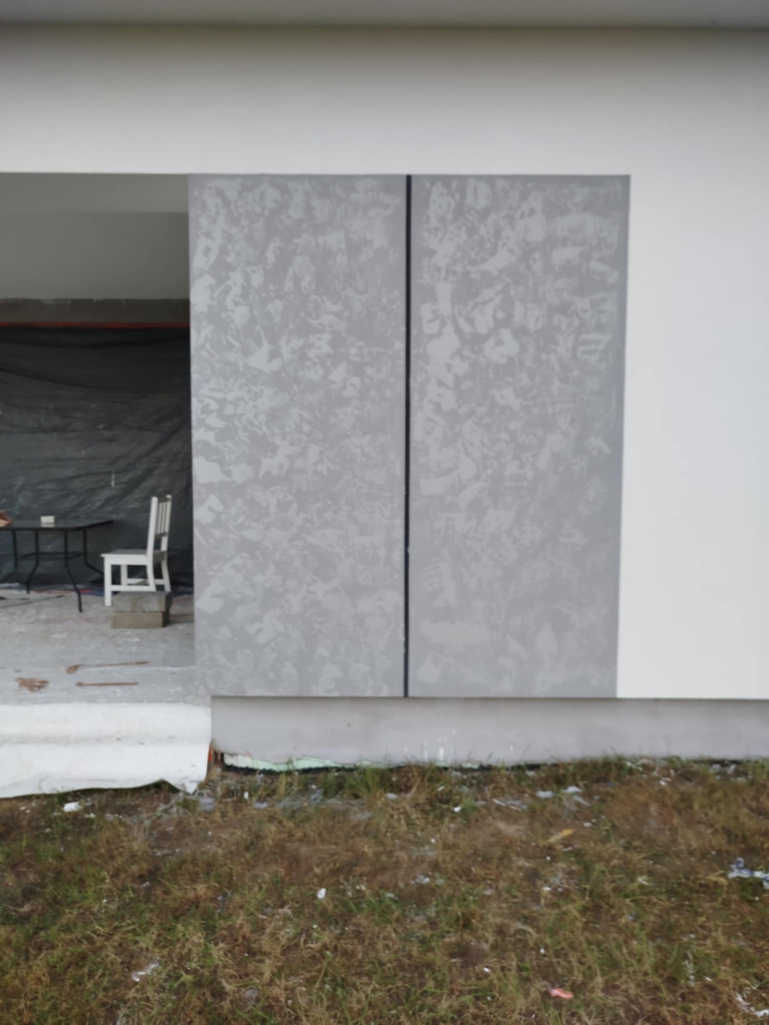 Wykończona elewacja budynku z szarą, dekoracyjną strukturą. W tle widoczne wnętrze z krzesłem i stołem pod przykryciem. Niski kadr z trawą na pierwszym planie.