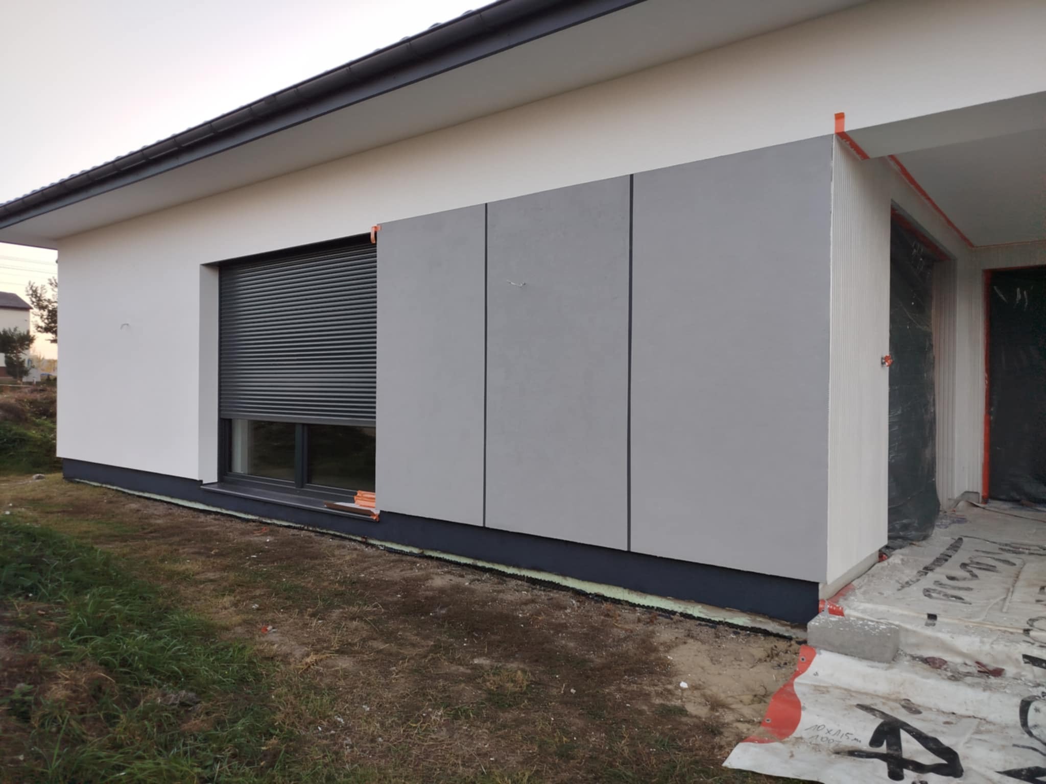 Elewacja domu jednorodzinnego z szarymi panelami elewacyjnymi, roletą okienną i przygotowanym wejściem. Widoczne detale wykończeniowe i materiały budowlane.