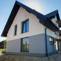 Dwukondygnacyjny dom z szarą dolną i białą górną elewacją, czarnym dachem i rynnami. Na bocznej ścianie rusztowanie. Błękitne niebo w tle, widok z dołu.