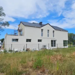 Dwukondygnacyjny dom z płaskim dachem w trakcie prac elewacyjnych; widoczne rusztowania i pracownik. Trawa na pierwszym planie. Błękitne niebo.