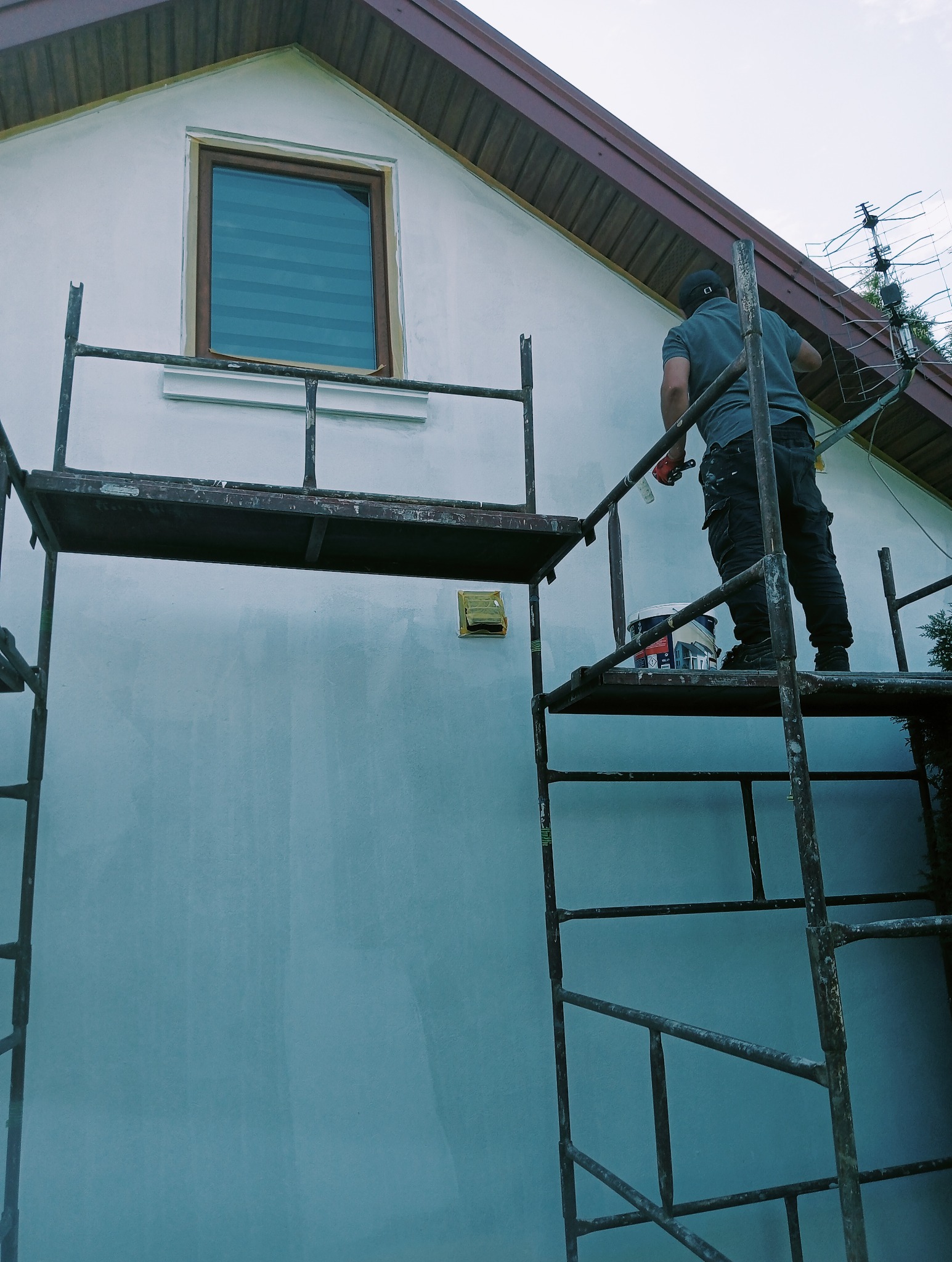 Mężczyzna na rusztowaniu maluje białą elewację domu jednorodzinnego, widoczny fragment okna z roletami, puszka z farbą i antena telewizyjna na dachu.