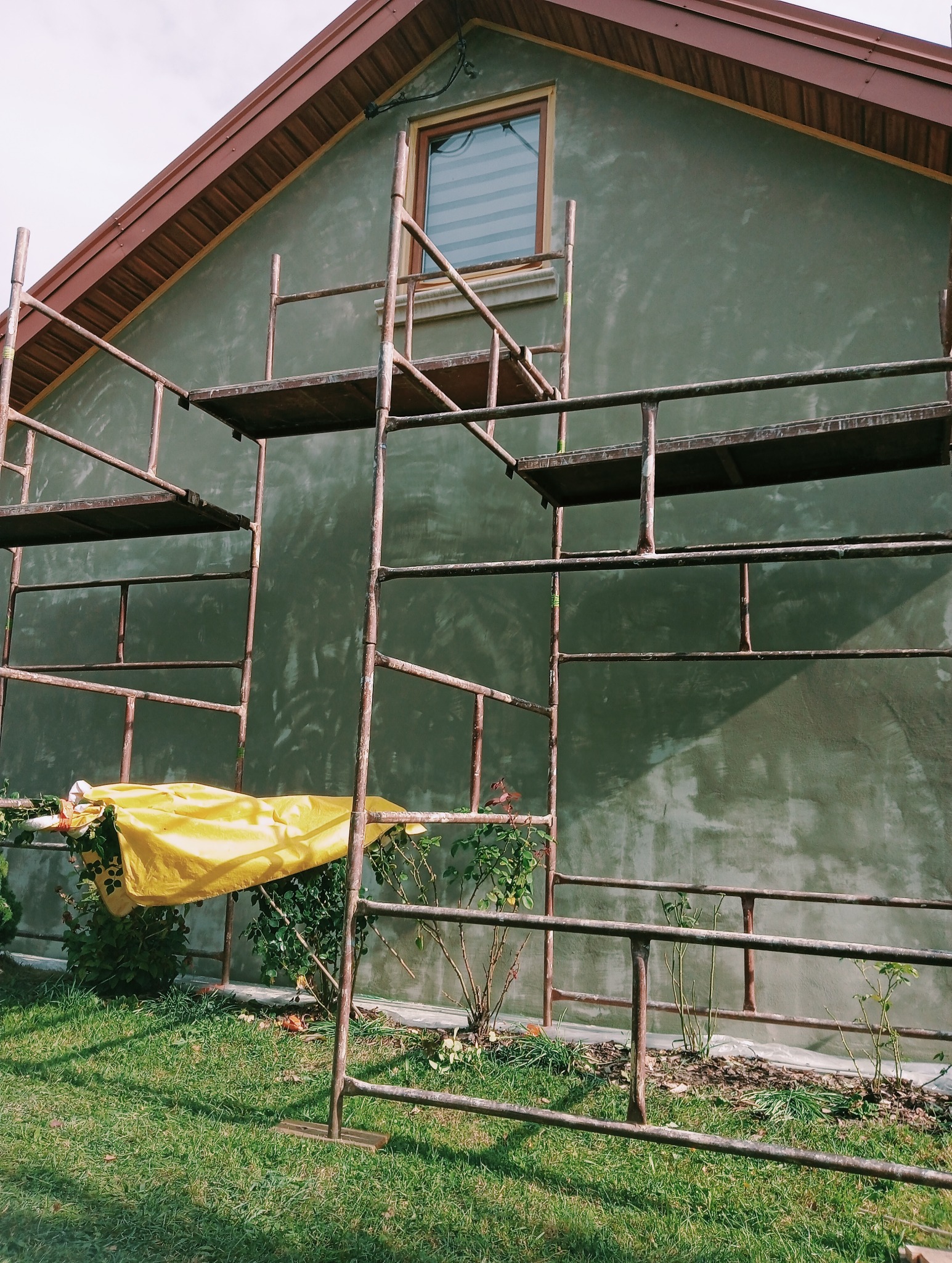 Dom w trakcie prac elewacyjnych, widoczne rusztowanie, fragment zielonego trawnika i krzewów, okno z żaluzjami, ściana pokryta szarym tynkiem, zabezpieczone rośliny żółtą folią.