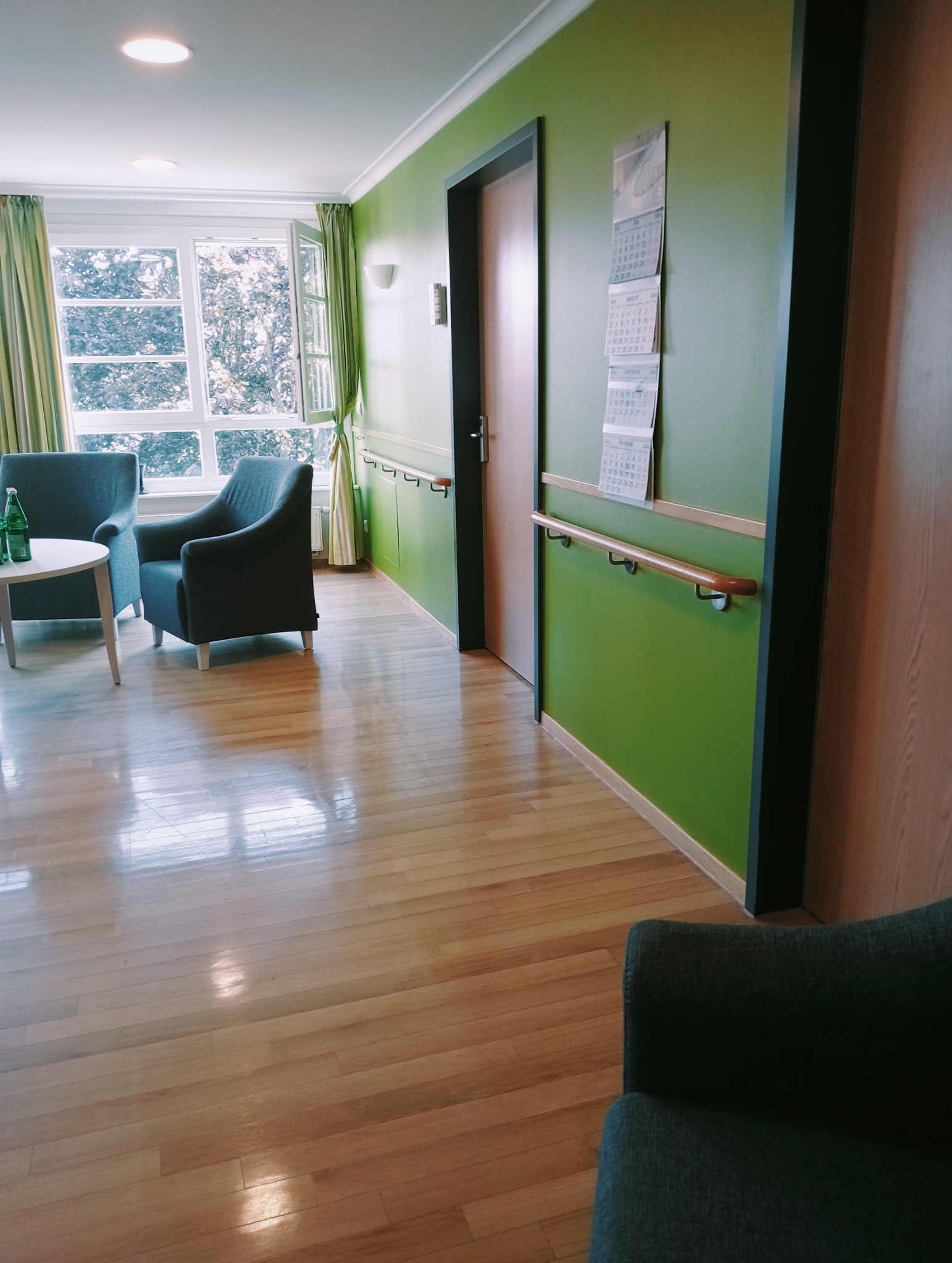 Jasne wnętrze pokoju z zielonymi ścianami, drewnianą podłogą i dwoma fotelami, widok na otwarte okno z firankami, kalendarze na ścianie i poręcze dla osób starszych.