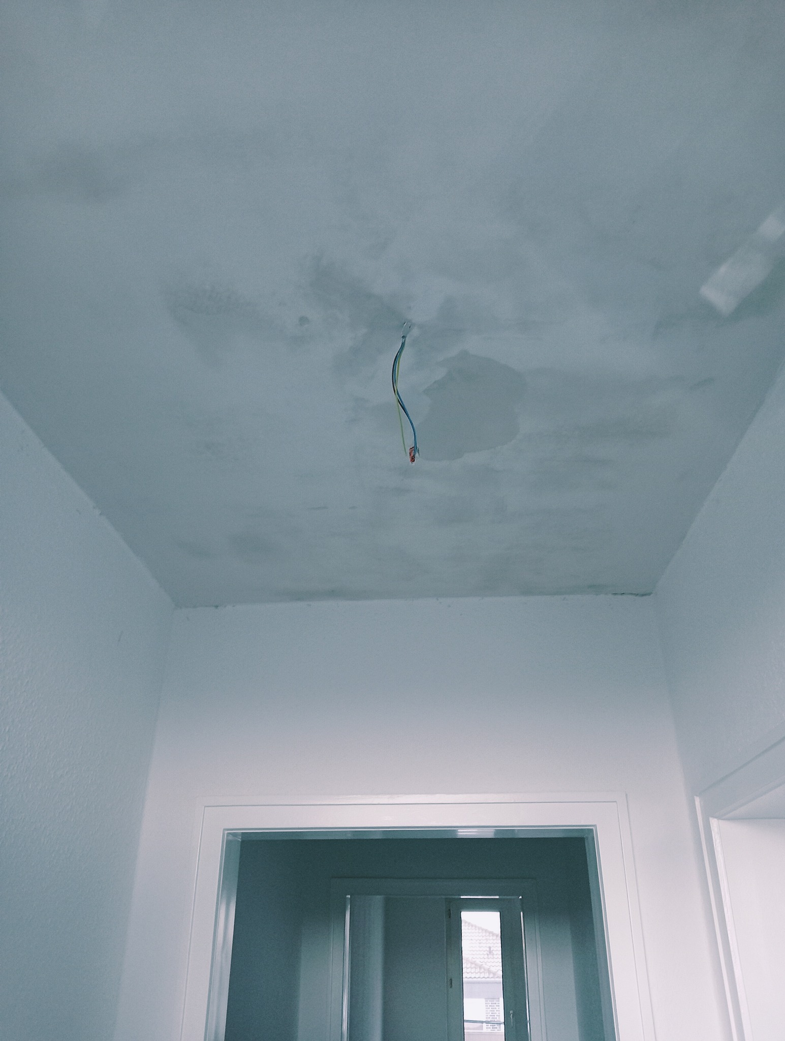 Surowy, niedokończony sufit w mieszkaniu z widocznymi śladami po malowaniu i wystającymi przewodami elektrycznymi, w tle widoczne drzwi do innego pomieszczenia.