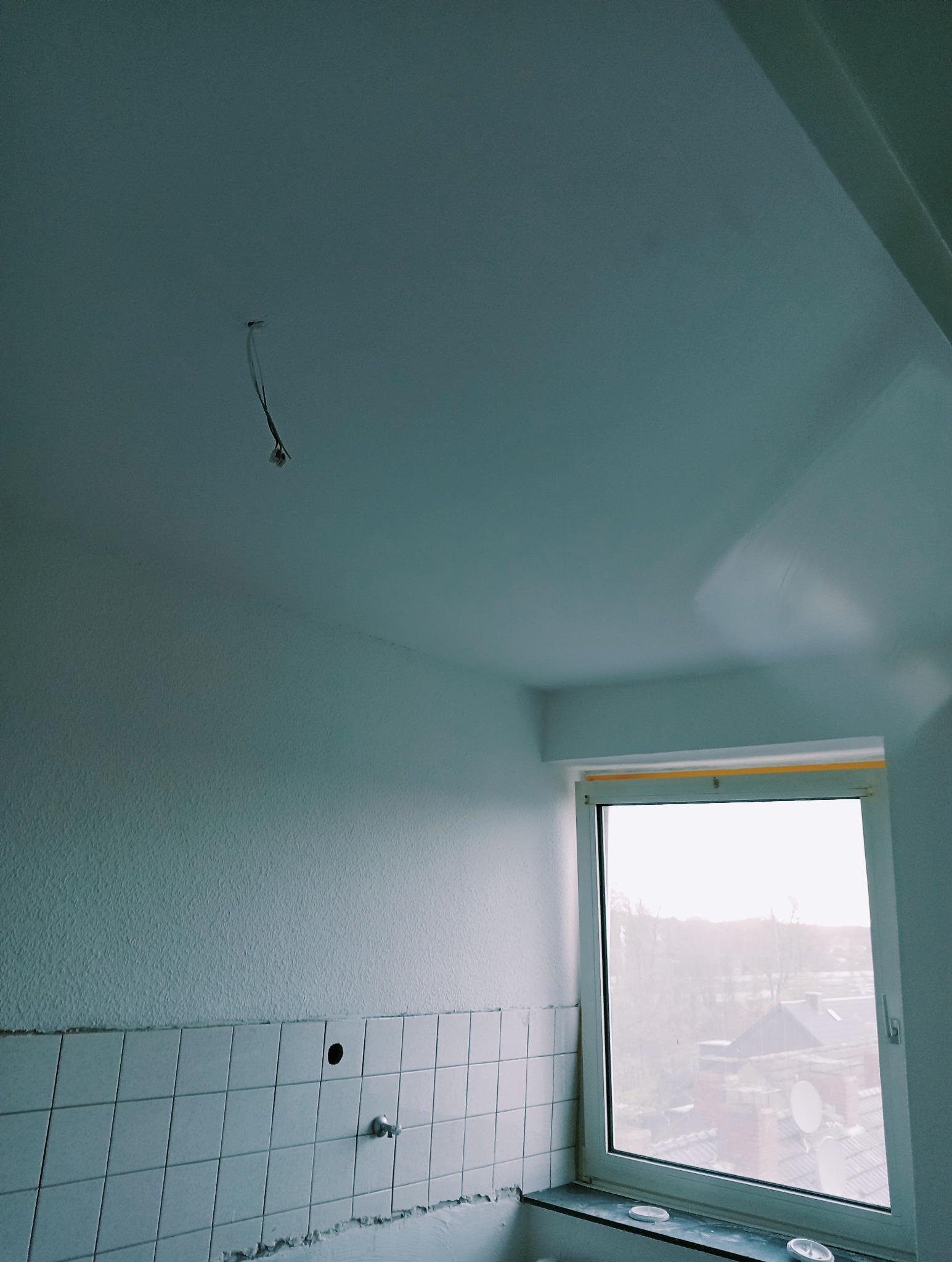 Pokój w trakcie malowania: świeżo pomalowany sufit na biało, ściany z fakturą, białe płytki na dole ściany, okno z widokiem na zewnątrz, wystające kable ze ściany.