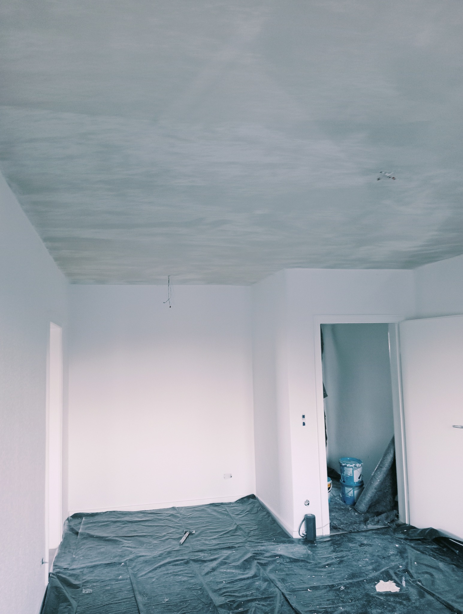 Przygotowane do malowania pomieszczenie z zabezpieczoną folią podłogą, świeżo pomalowanym na biało sufitem i ścianami, widoczne puszki z farbą w sąsiednim pokoju.