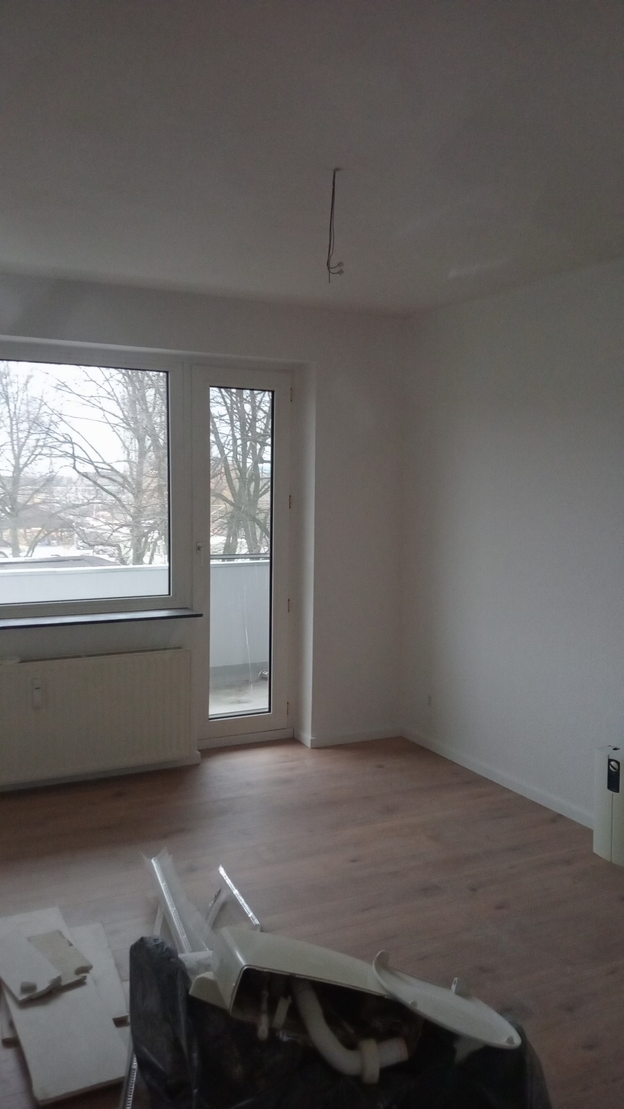 Pusty pokój z nową, brązową podłogą panelową, białe ściany, okno balkonowe z widokiem na drzewa zimą, na podłodze leżą resztki po montażu paneli.