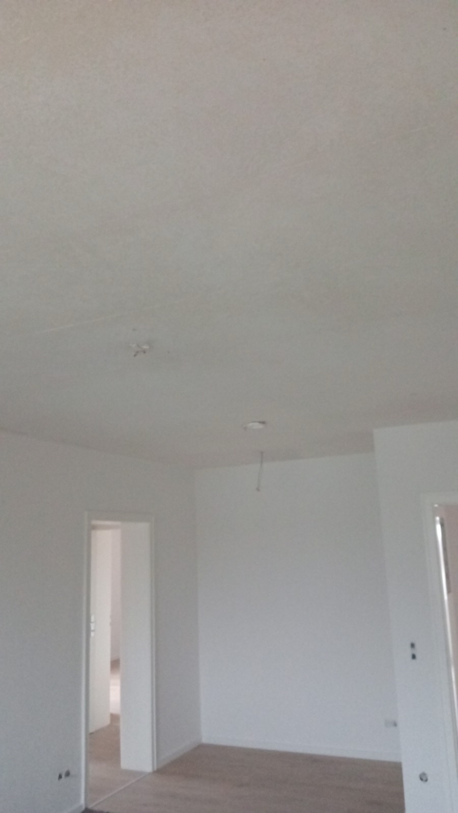 Wnętrze pokoju po malowaniu ścian na biało; widoczne gładkie ściany, sufit z niedoskonałościami i fragment podłogi z panelami.
