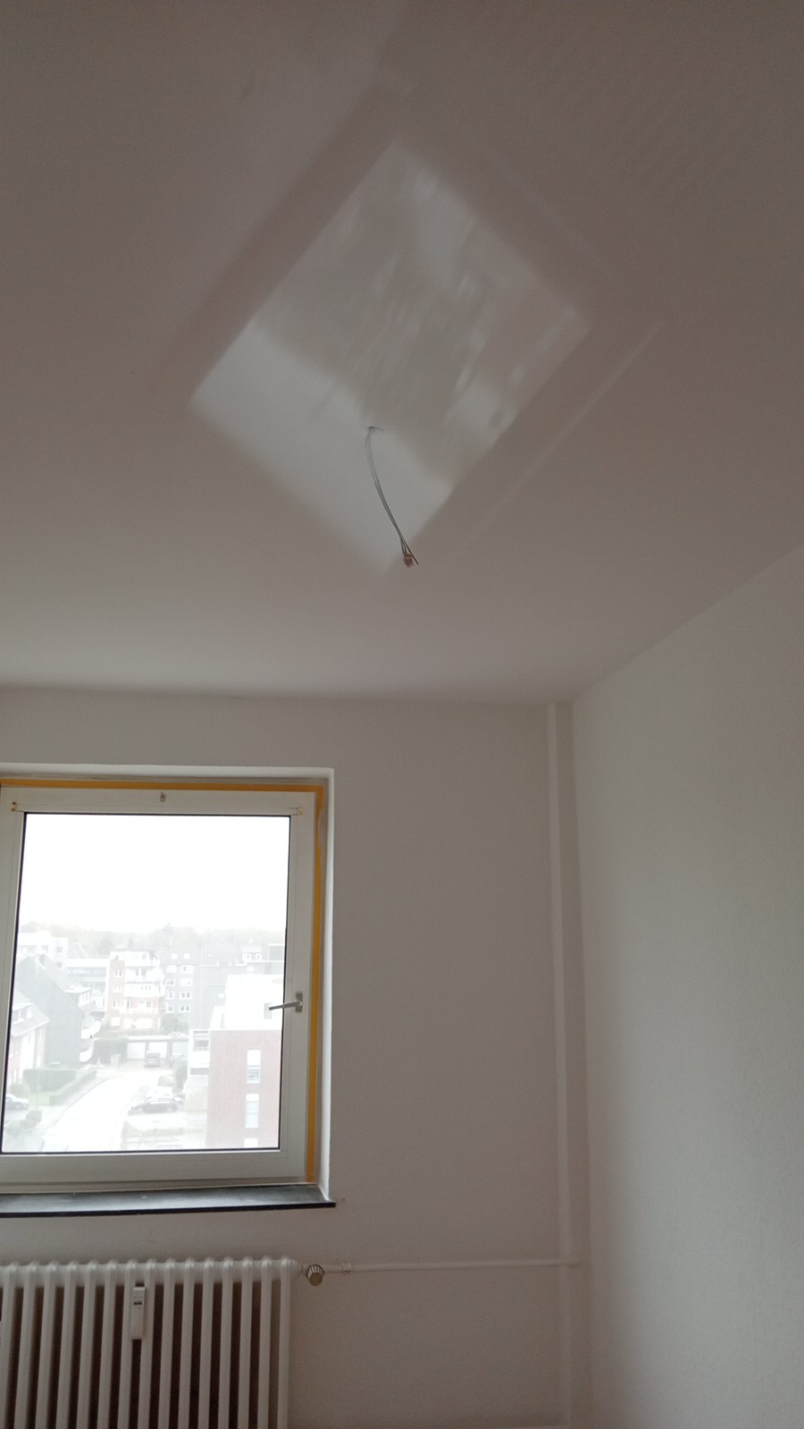 Białe wnętrze pokoju z widocznym oknem, grzejnikiem i instalacją elektryczną wystającą z otworu w suficie, przygotowane do dalszych prac wykończeniowych.