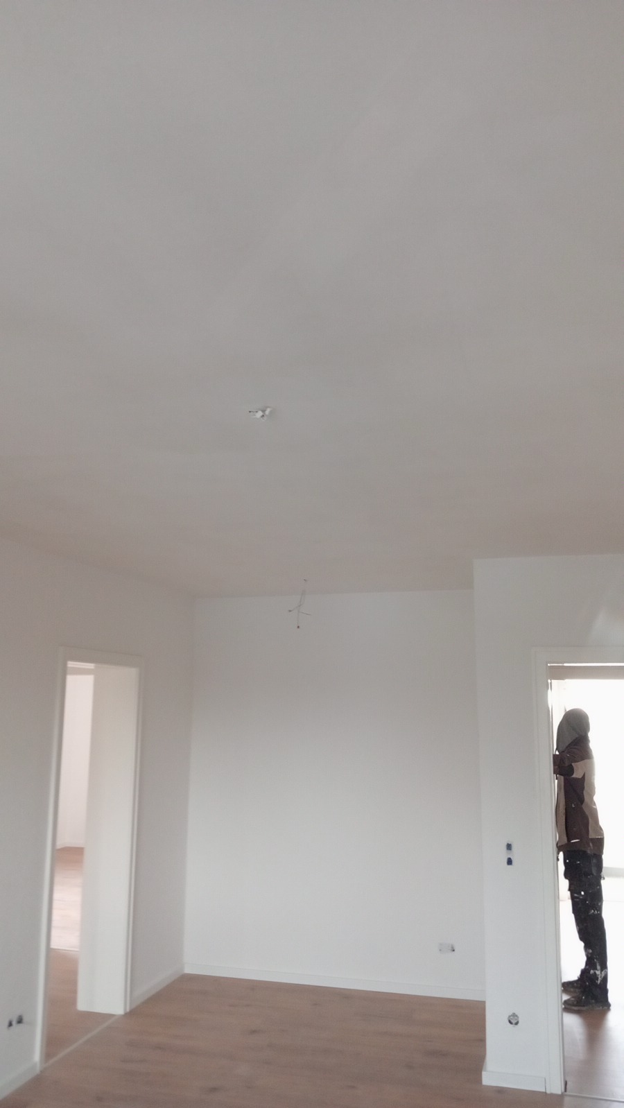 Wnętrze świeżo pomalowanego na biało pomieszczenia z drewnianą podłogą; malarz w roboczym ubraniu widoczny w przejściu drzwiowym po prawej stronie.