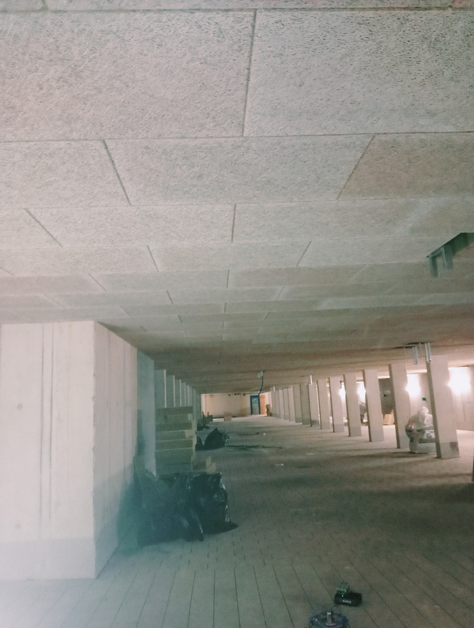 Wnętrze w trakcie prac wykończeniowych, widoczne białe ściany, sufit z płyt o fakturze drewna, podłoga z drewnianych desek, czarne worki na śmieci i narzędzia na podłodze.