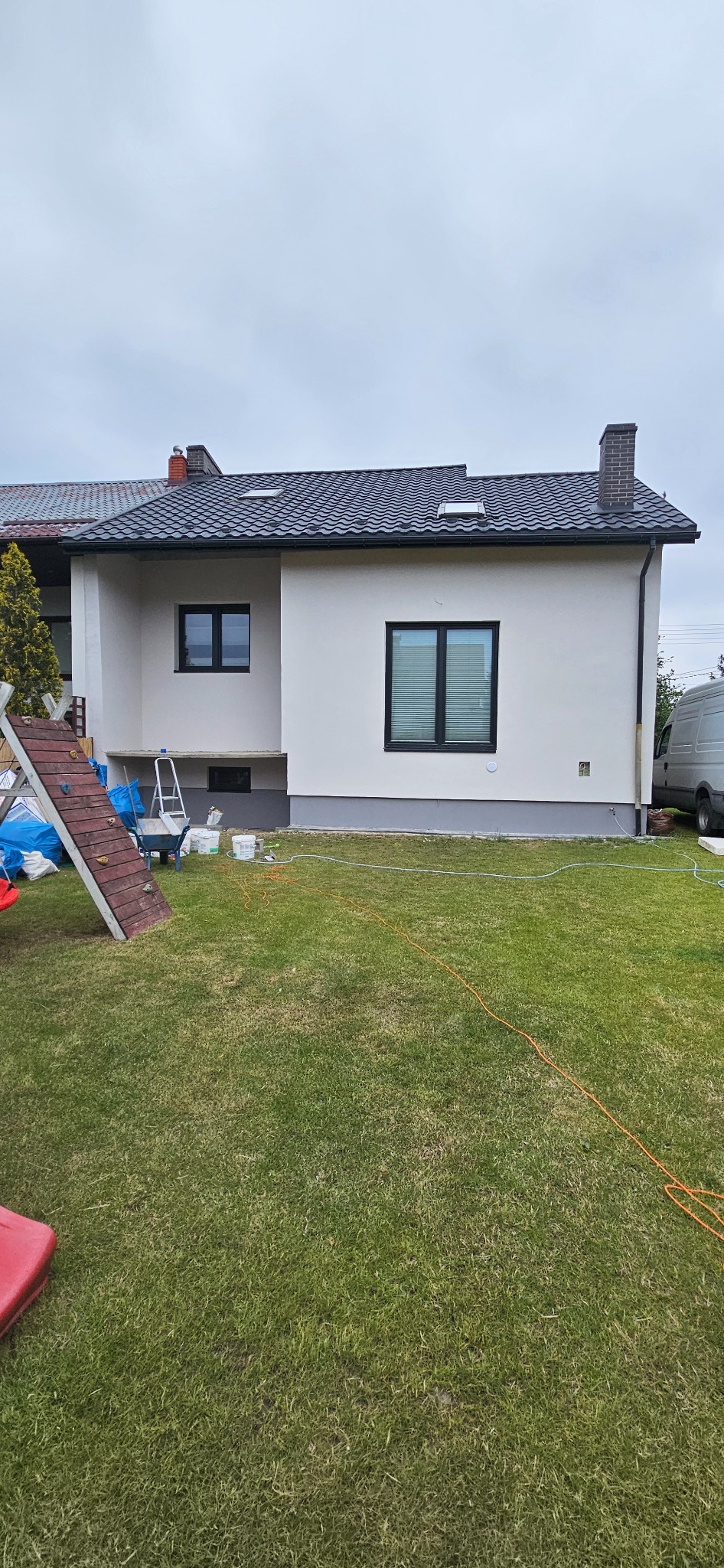 Biała elewacja domu jednorodzinnego po renowacji, widok z ogrodu. Widoczne okna z czarnymi ramami, fragment drabiny i narzędzia malarskie.