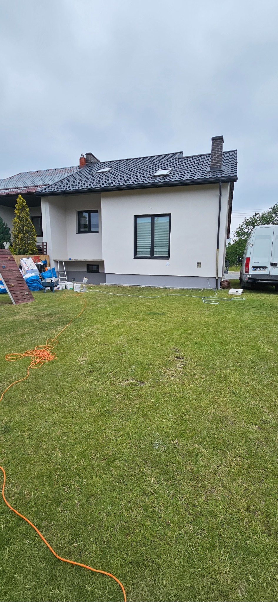 Biała elewacja domu jednorodzinnego z czarnymi oknami, widok z trawnika, przygotowane materiały budowlane, drabina, częściowo widoczny samochód dostawczy.