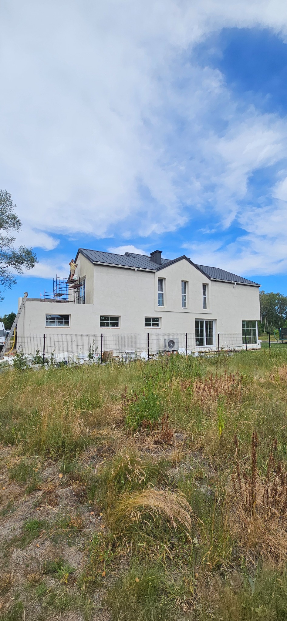 Dwukondygnacyjny dom z płaskim dachem w trakcie prac elewacyjnych; widoczne rusztowania i pracownik. Trawa na pierwszym planie. Błękitne niebo.