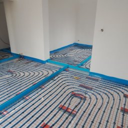 HydroExpert Adam Pisarek - Niedokończona instalacja ogrzewania podłogowego w nowym budynku z widocznymi niebieskimi rurami na siatce i folii, z niebieską taśmą przy ścianach.