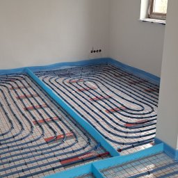 HydroExpert Adam Pisarek - Niedokończona instalacja ogrzewania podłogowego w nowym budownictwie, widoczne rury grzewcze w kształcie serpentyny przymocowane do siatki z izolacją, nieotynkowane ściany z wystającymi kablami...
