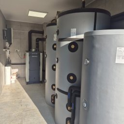HydroExpert Adam Pisarek - Kotłownia z piecem RAKOCZY i trzema szarymi zbiornikami akumulacyjnymi, połączonymi czarnymi rurami. Na ścianie widoczny panel sterowania. Pomieszczenie wyłożone jasnymi płytkami.