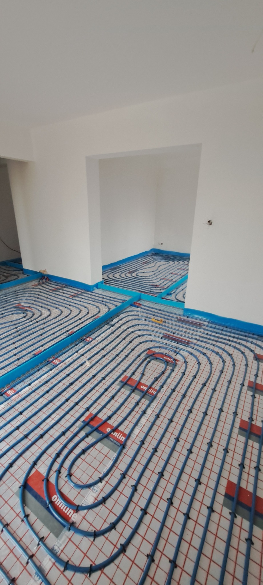 Niedokończona instalacja ogrzewania podłogowego w nowym budynku z widocznymi niebieskimi rurami na siatce i folii, z niebieską taśmą przy ścianach.