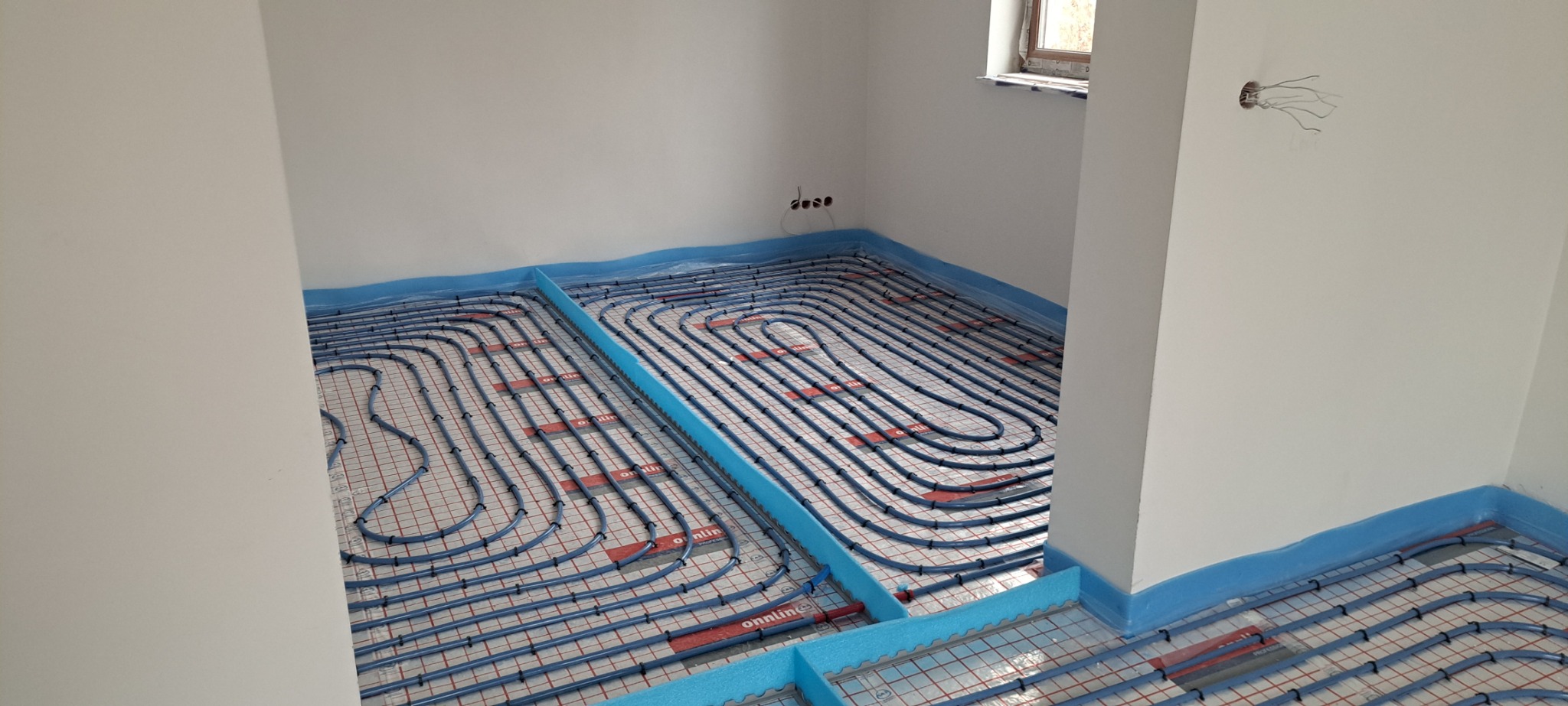 Niedokończona instalacja ogrzewania podłogowego w nowym budownictwie, widoczne rury grzewcze w kształcie serpentyny przymocowane do siatki z izolacją, nieotynkowane ściany z wystającymi kablami...